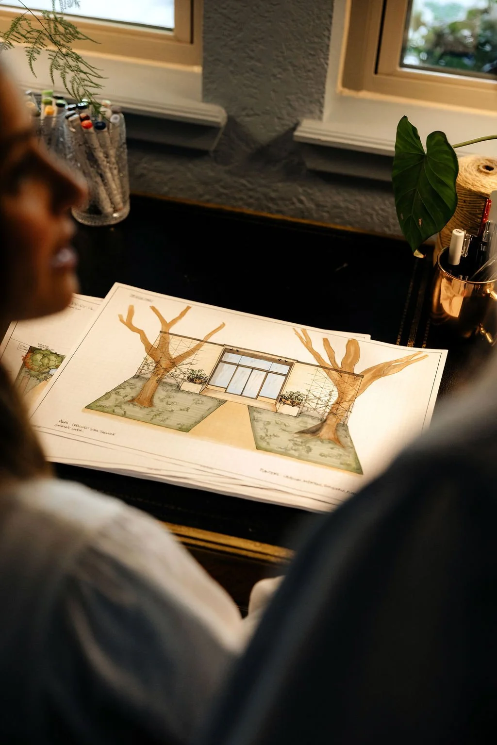 Design drawing of a building entrance with large glass doors between two trees, shown on a desk with plants and office supplies nearby.