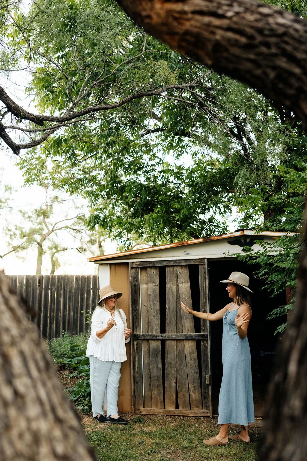 Two women are having a conversation outside near a small wooden shed, surrounded by trees. One woman is wearing a white outfit and a wide-brimmed hat, while the other is in a light blue dress and a wide-brimmed hat, gesturing with her hands.