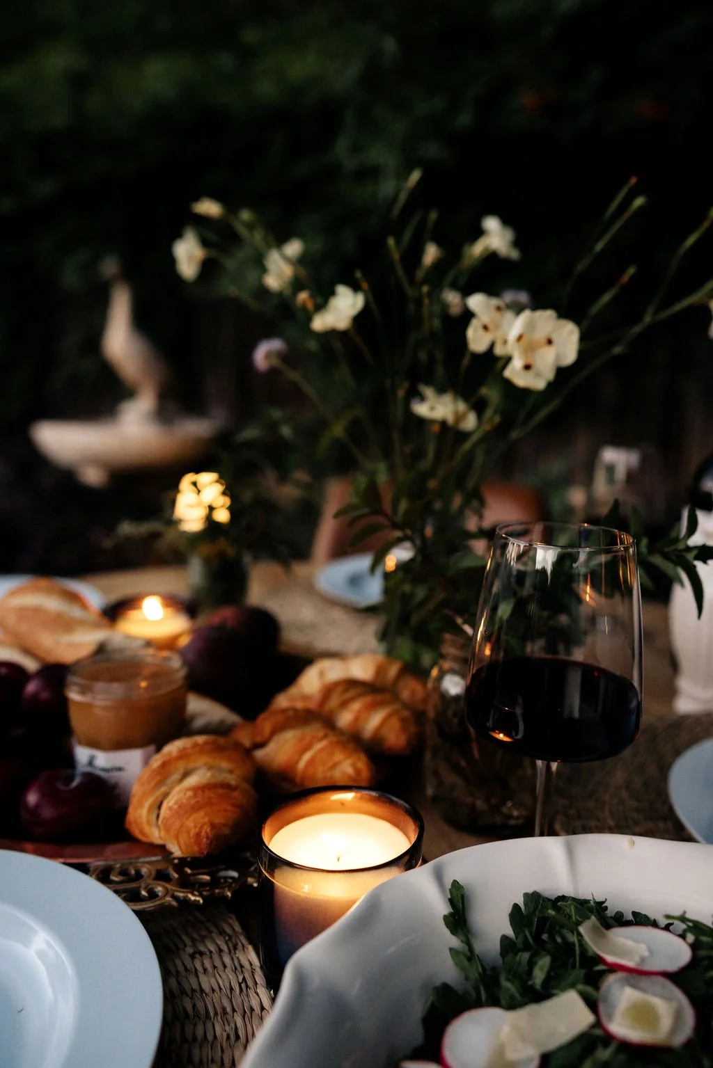 A candlelit dinner table with a glass of red wine, croissants, a jar of jam, fresh grapes, a salad with radishes, and a floral centerpiece featuring white flowers.