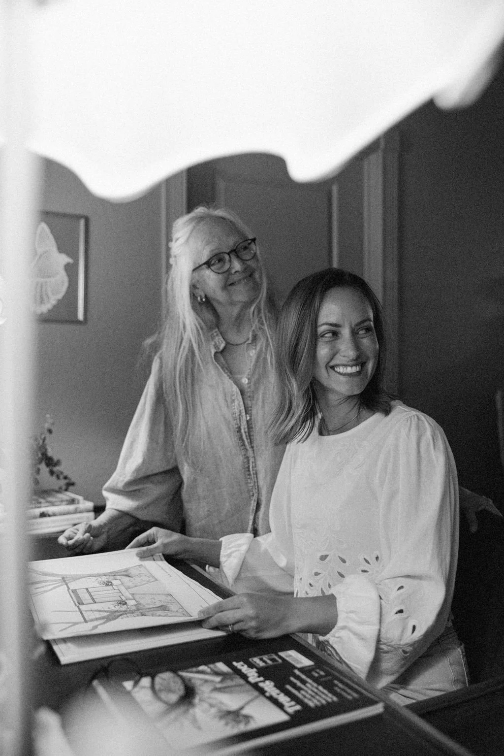 Two women, one older with glasses and long hair, the other younger with shoulder-length hair, smiling and looking at each other while looking at an open photo album or book on a desk, in a room with framed pictures on the wall.