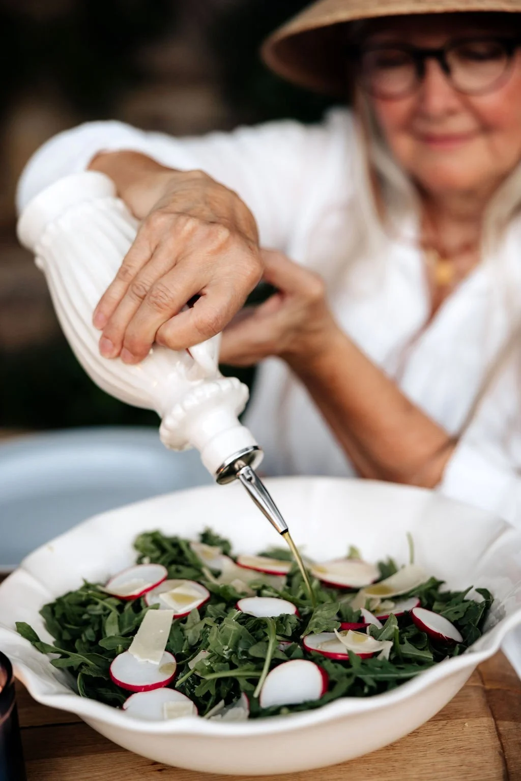 Older woman wearing a white shirt, straw hat, and glasses pouring oil over a fresh arugula salad with radish slices and shaved cheese in a white ceramic bowl.