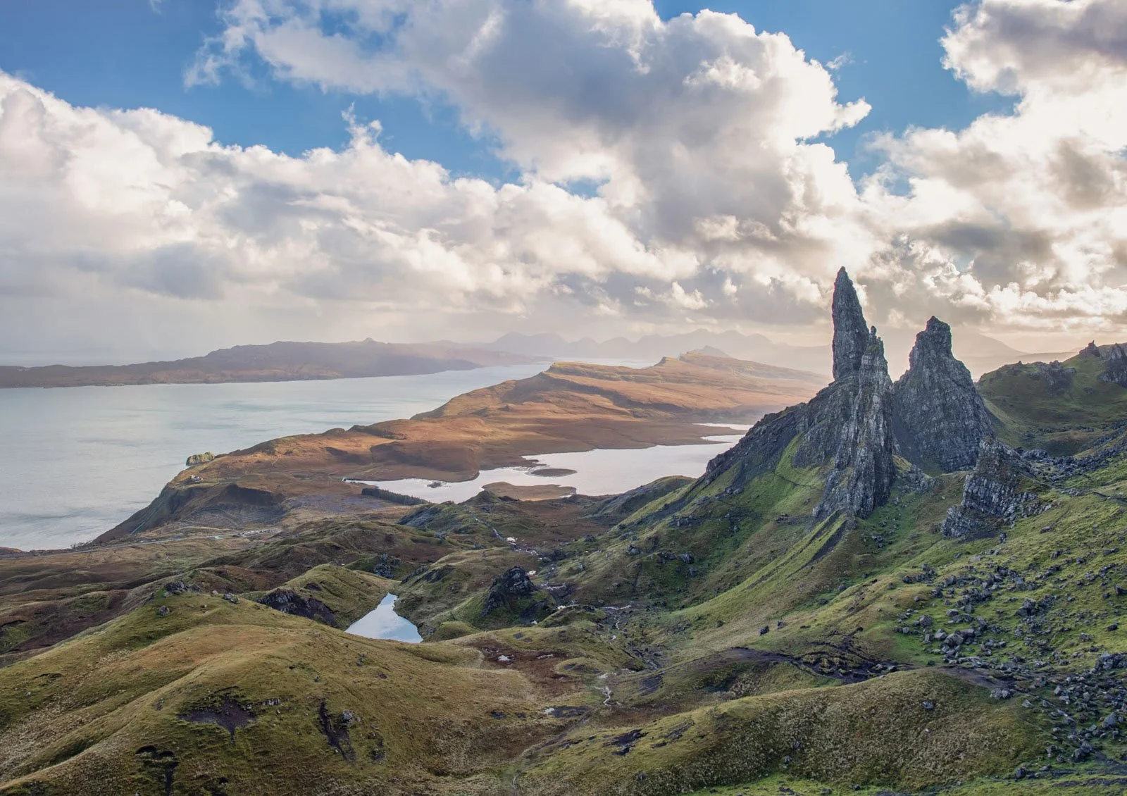 A scenic landscape featuring green rolling hills, rocky formations, a body of water, and a partly cloudy sky.