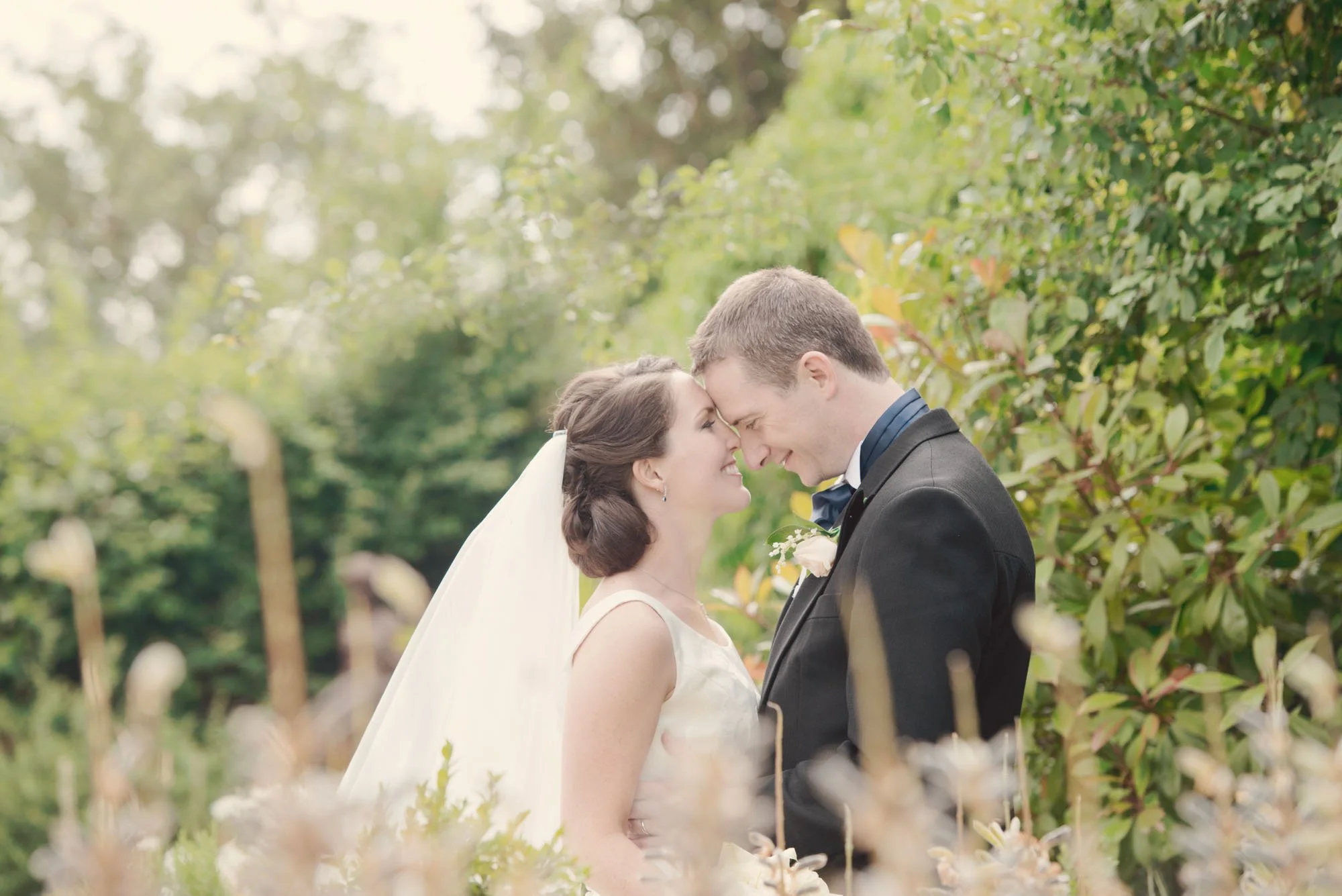 A bride and groom standing close together in the garden with foreheads touching during their wedding celebration at Northbrook Park in Surrey