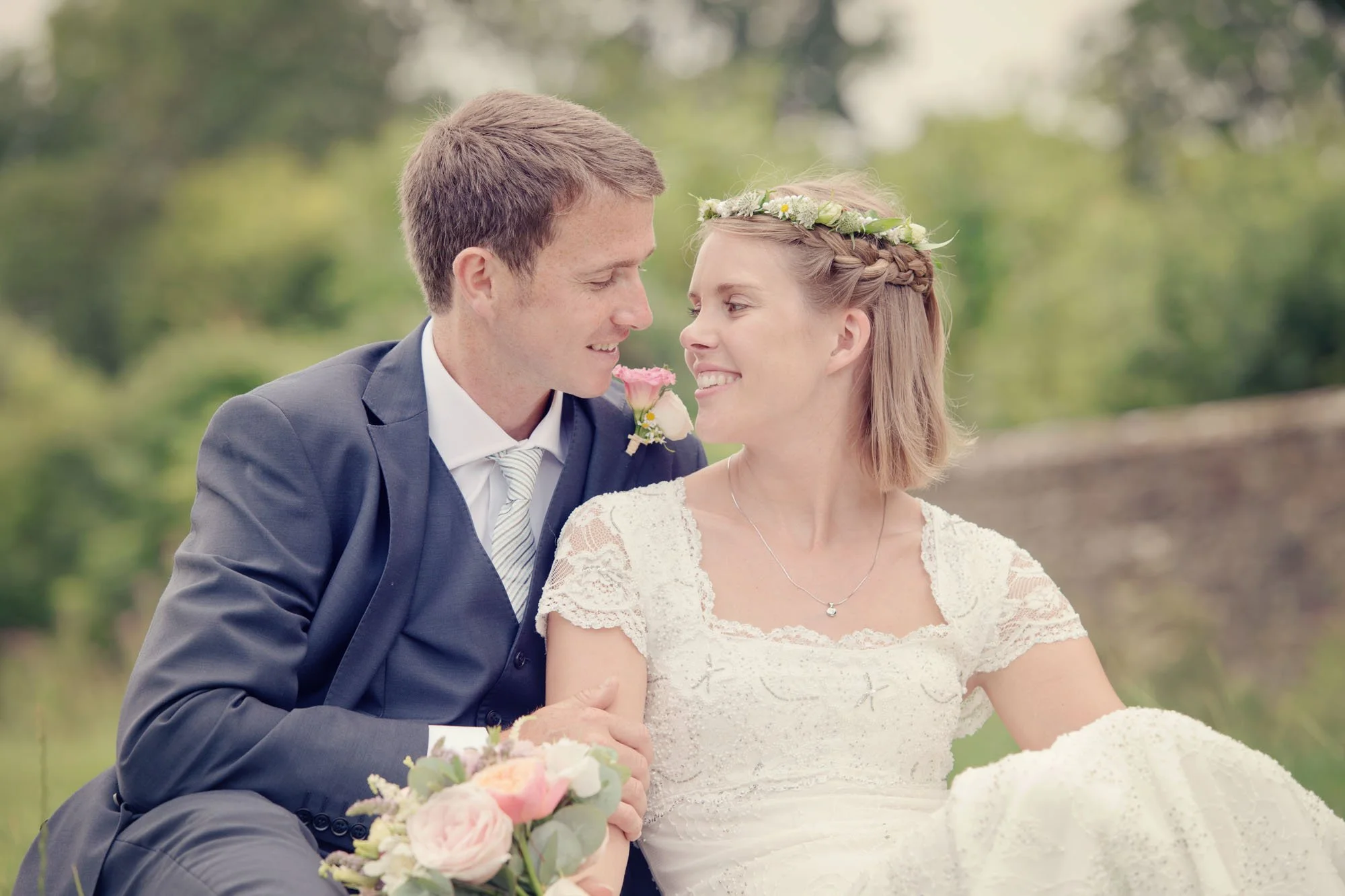A bride and groom sit on the grass in the walled garden in Kenwood House in Hampstead Heath, north London.  The bride wears a ring of flowers in her hair and holds a bouquet, and the bride and groom gaze at each other smiling lovingly.  
