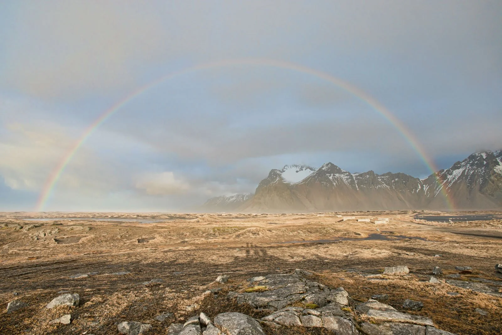 A landscape with a double rainbow over rocky terrain and snow-capped mountains in the distance.
