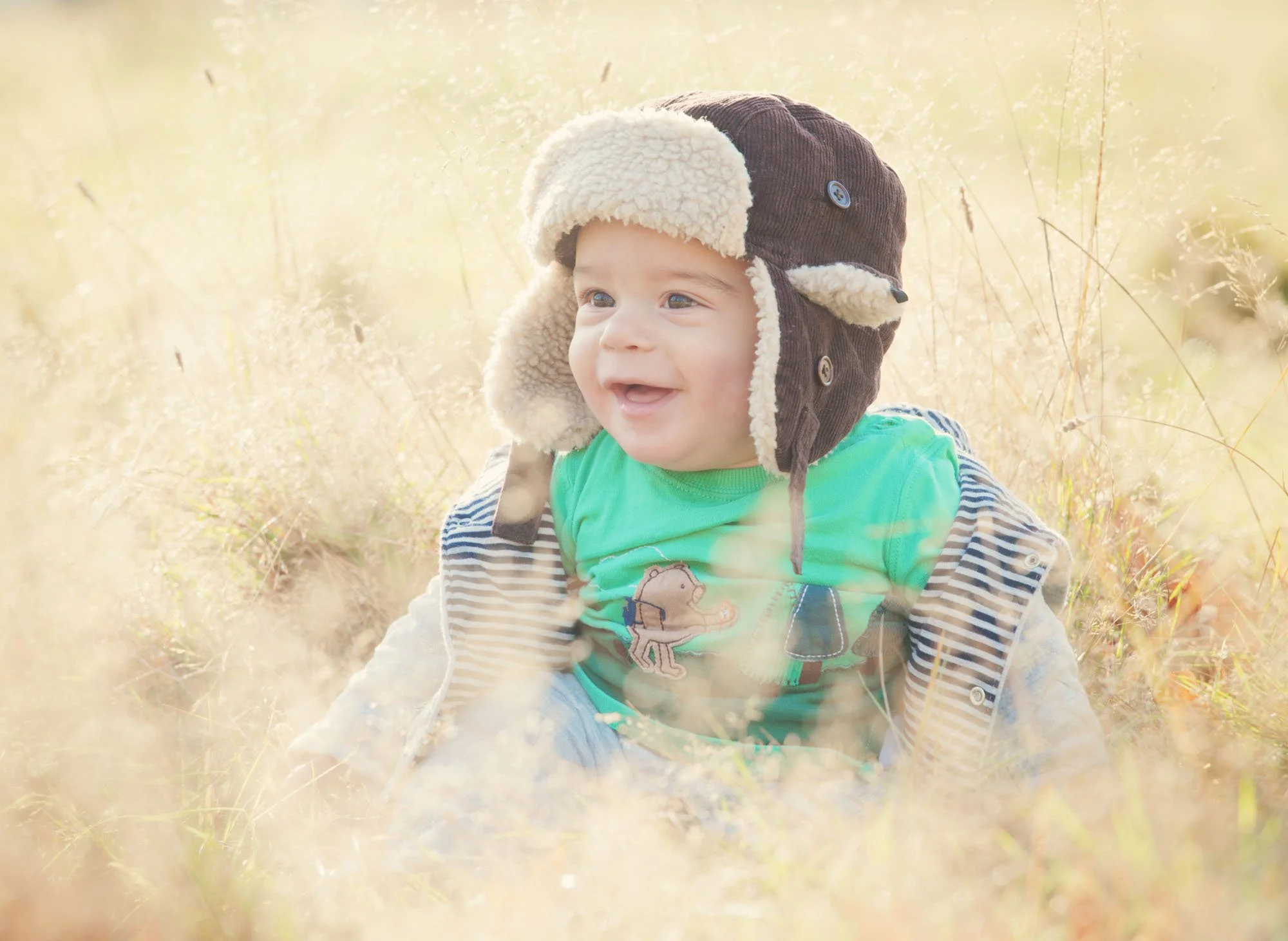 Eight month old baby boy wears a sheepskin aviator hat with earflaps as he sits smiling in the long grass during a family photoshoot in Hampstead Heath.  He is surrounded by golden light.