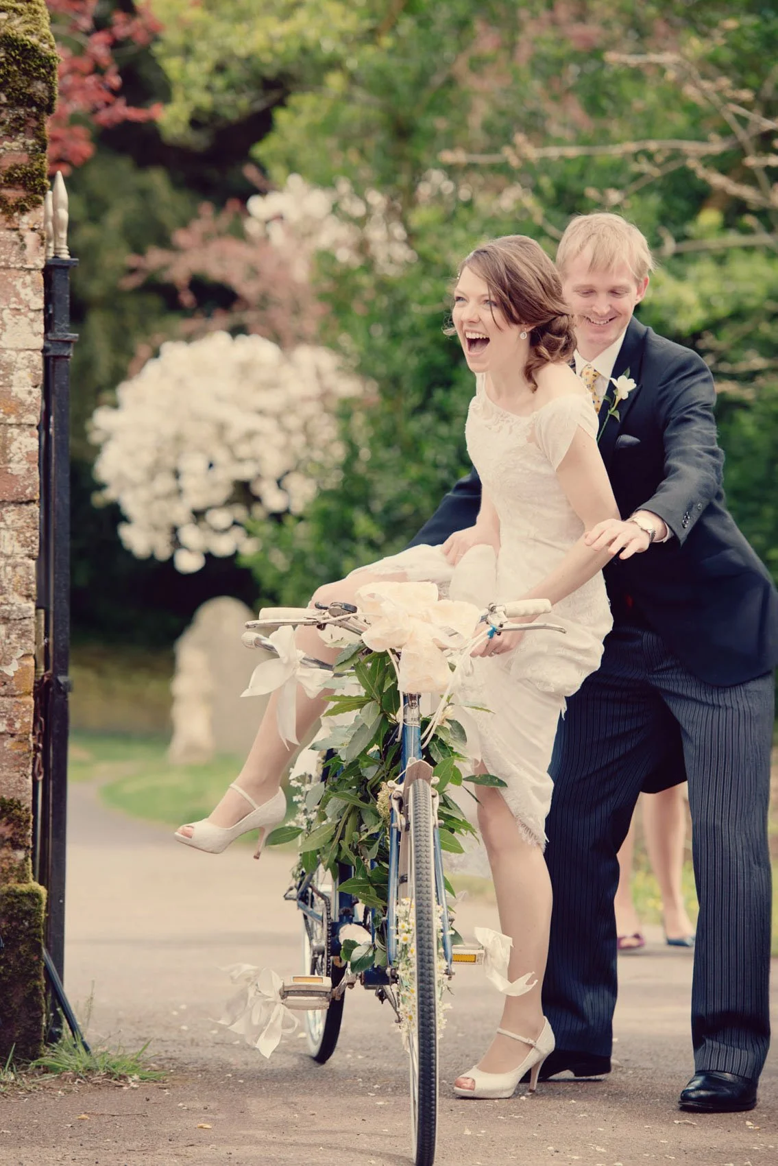 A groomsman helps a laughing bride up onto a bicycle decorated with flowers, during a spring wedding in Surrey