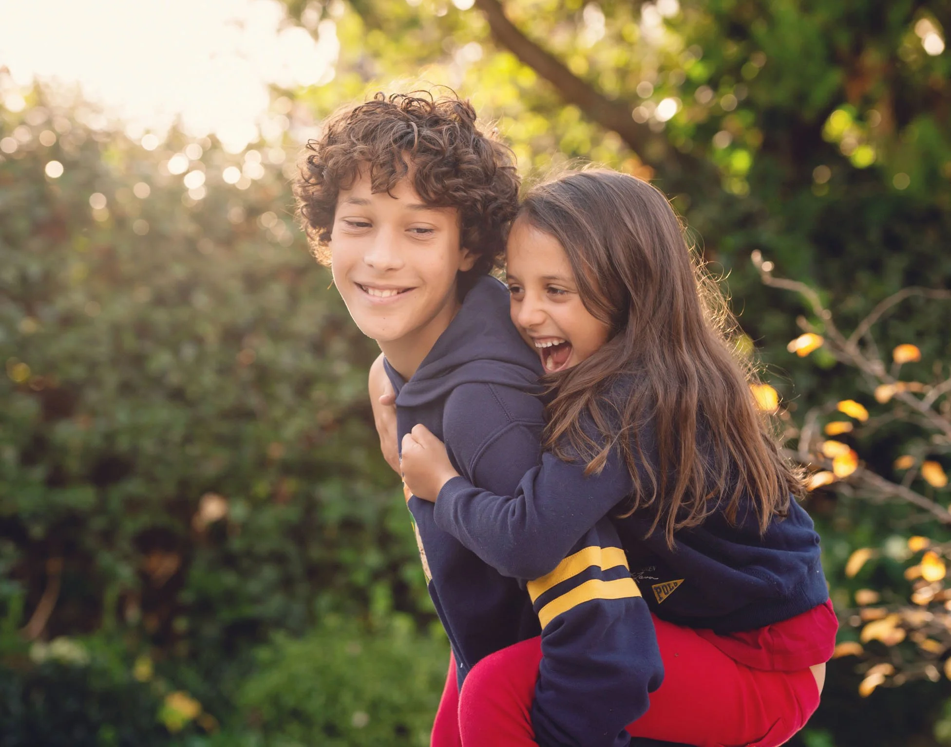 A young teenage boy carries his little sister on his back and both smile in the golden winter light, during a family photoshoot in Notting Hill in west London 