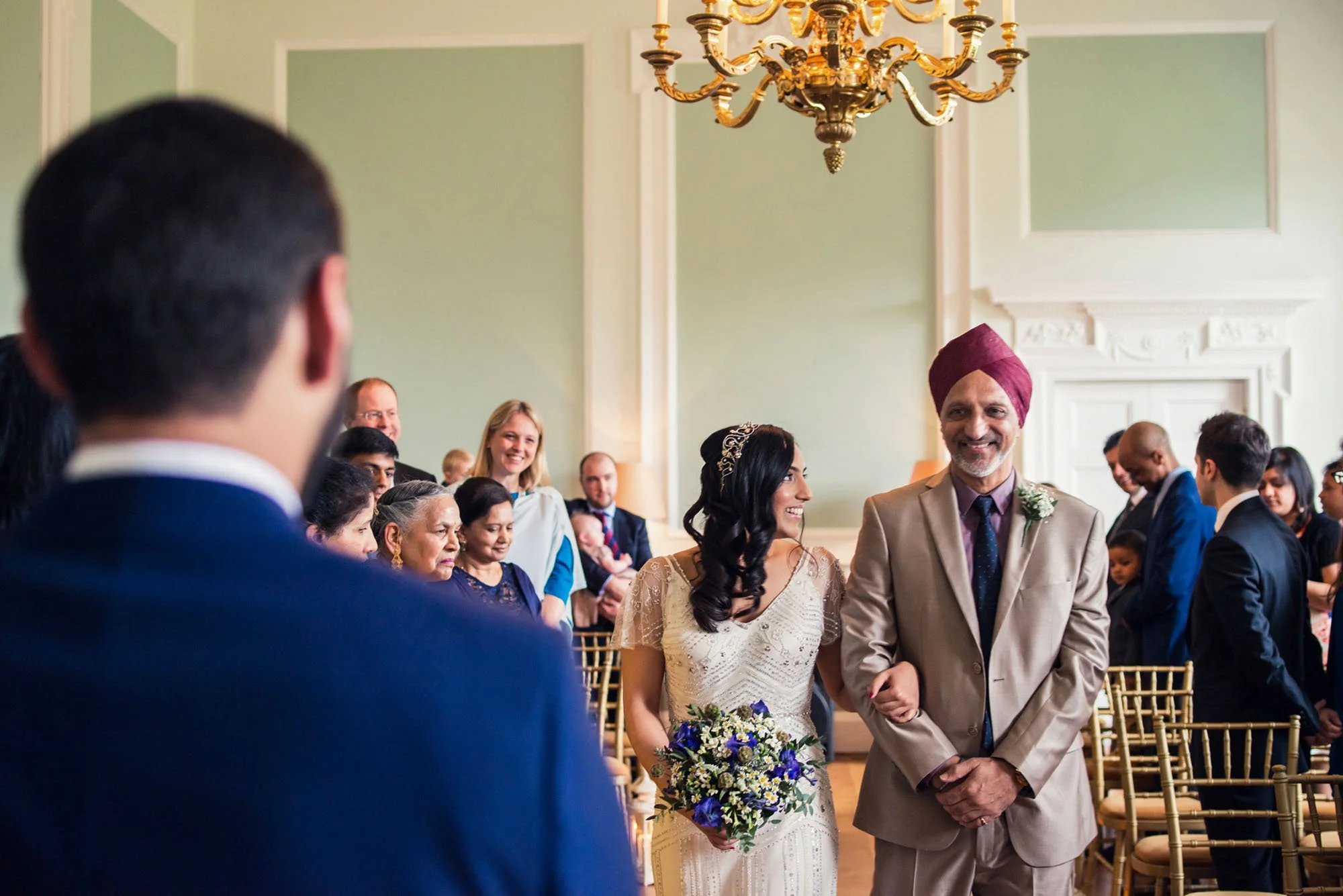 A bride and her father walk down the aisle at a wedding ceremony at Botleys Mansion in Surrey, as the bride's father smiles as the waiting groom