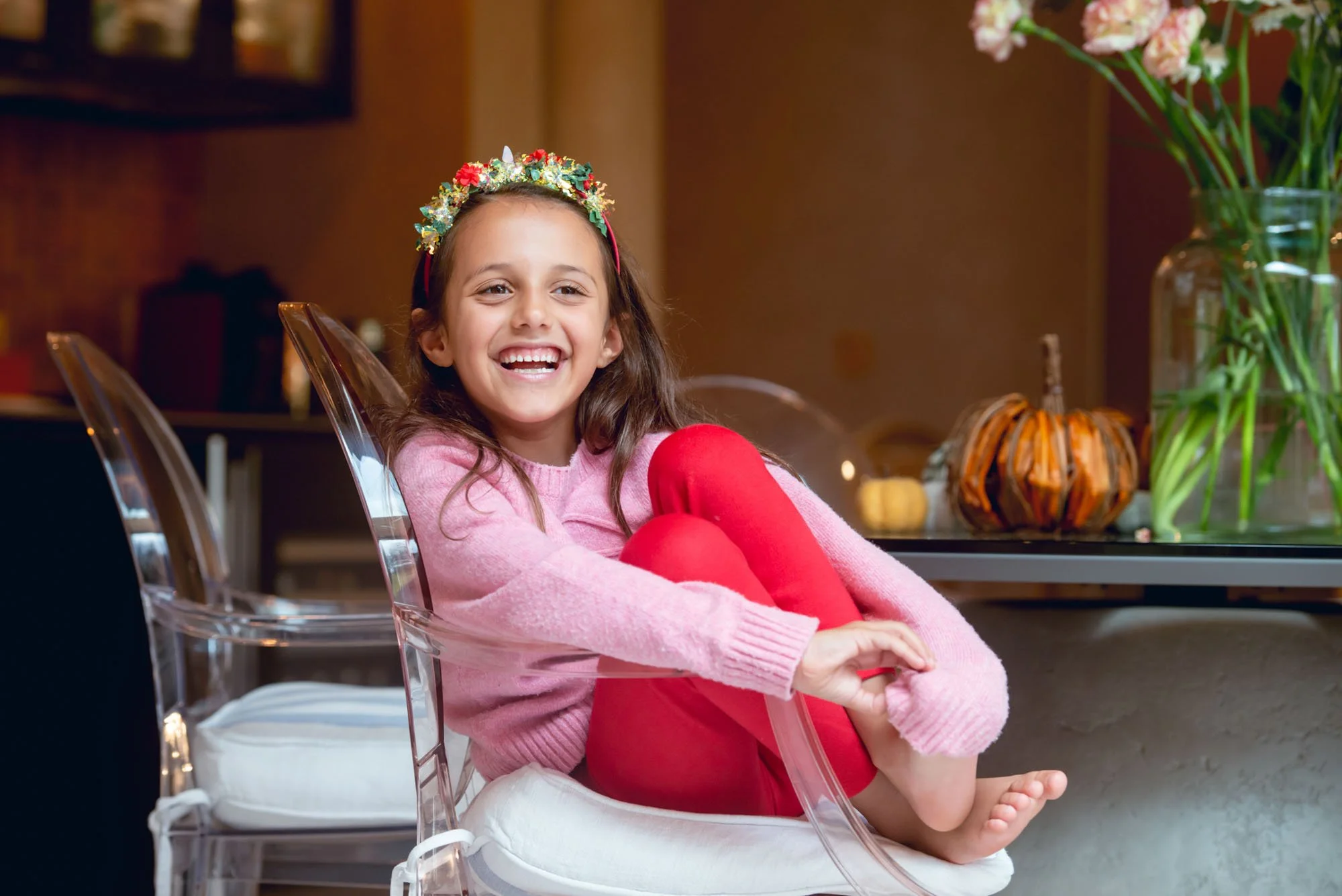 A little girl wearing a sparkly colourful headband sits laughing on a dining chair at her home in Hampstead in north London