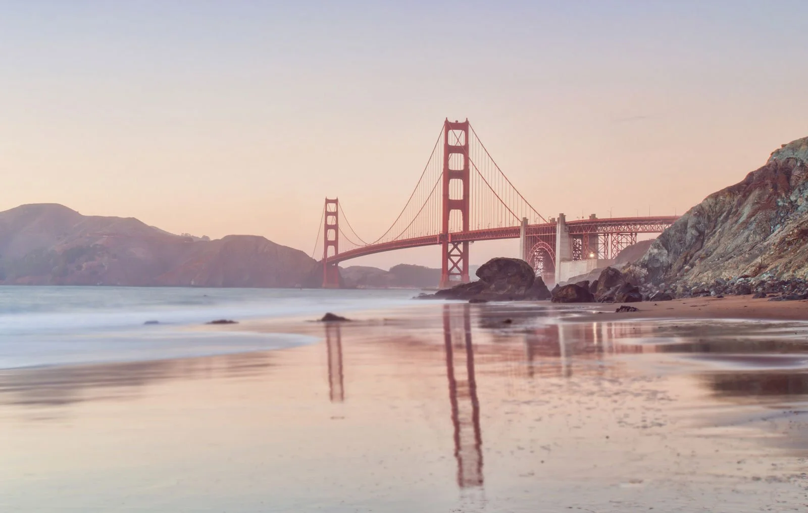 A sunrise or sunset view of the Golden Gate Bridge in San Francisco, California, with calm water and rocky shores in the foreground.
