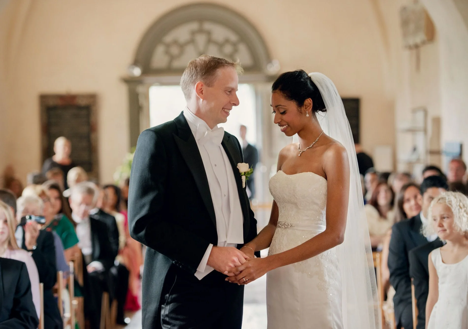 A new bride smiles shyly down as she holds the hands of her proud new husband at their family chapel in Hertfordshire