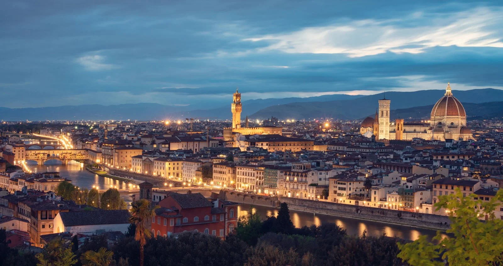 A panoramic view of Florence, Italy at dusk, featuring the Florence Cathedral with its dome, the Palazzo Vecchio with its tower, and the Arno River reflecting city lights, with mountains in the background.