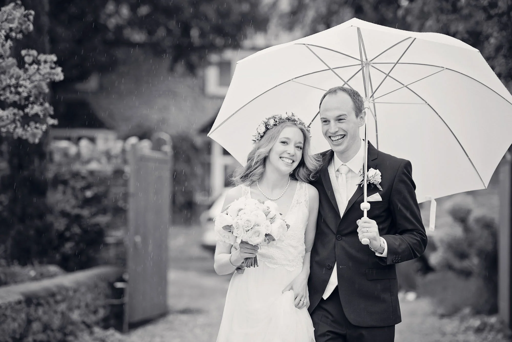 A bride and groom smile as they walk under a white umbrella in the rain, following their micro wedding in St Albans in Hertfordshire