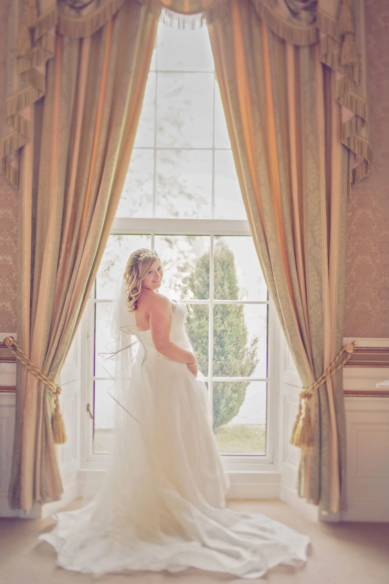 A bride stands in front of a full length window with curtains during her micro wedding at her family home in Surrey