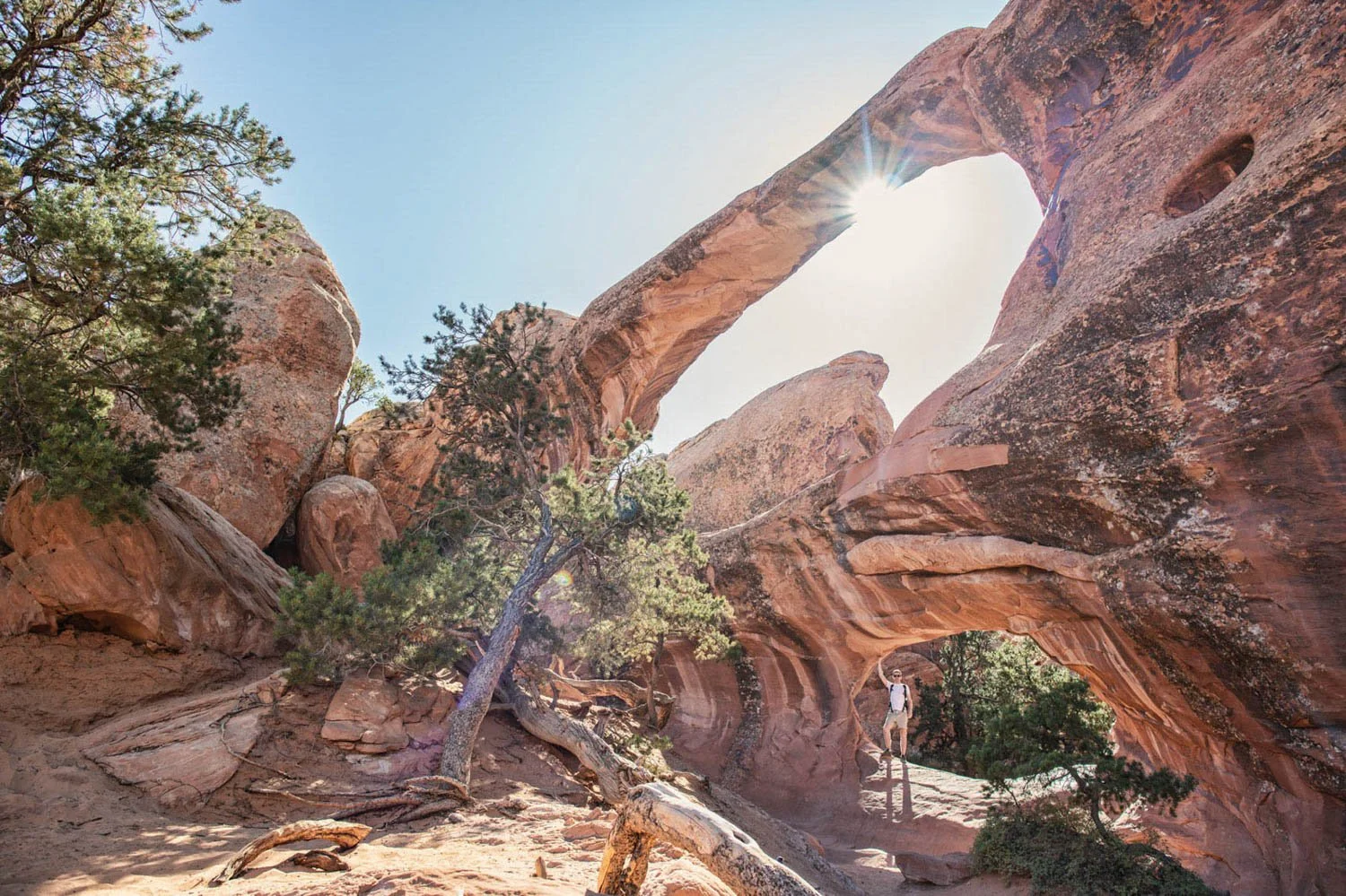 A person hiking through a desert landscape with large red rock formations and natural arches, with the sun shining through one of the arches.