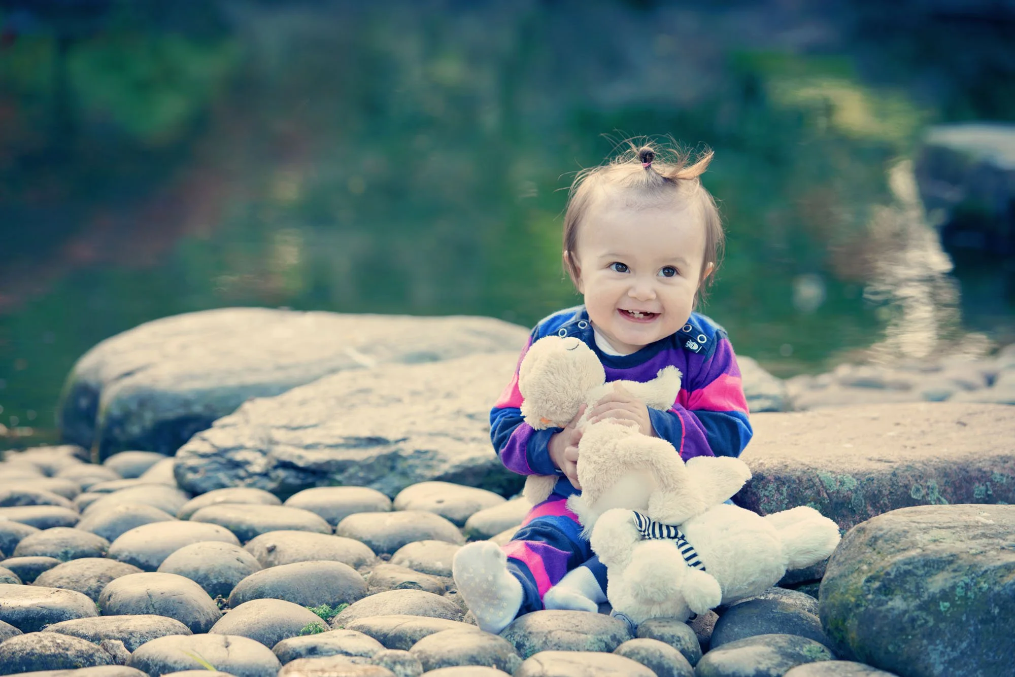 Little girl cuddles a soft toy and laughs as she sits on pebbles next to a pond during a family photoshoot in Holland Park London