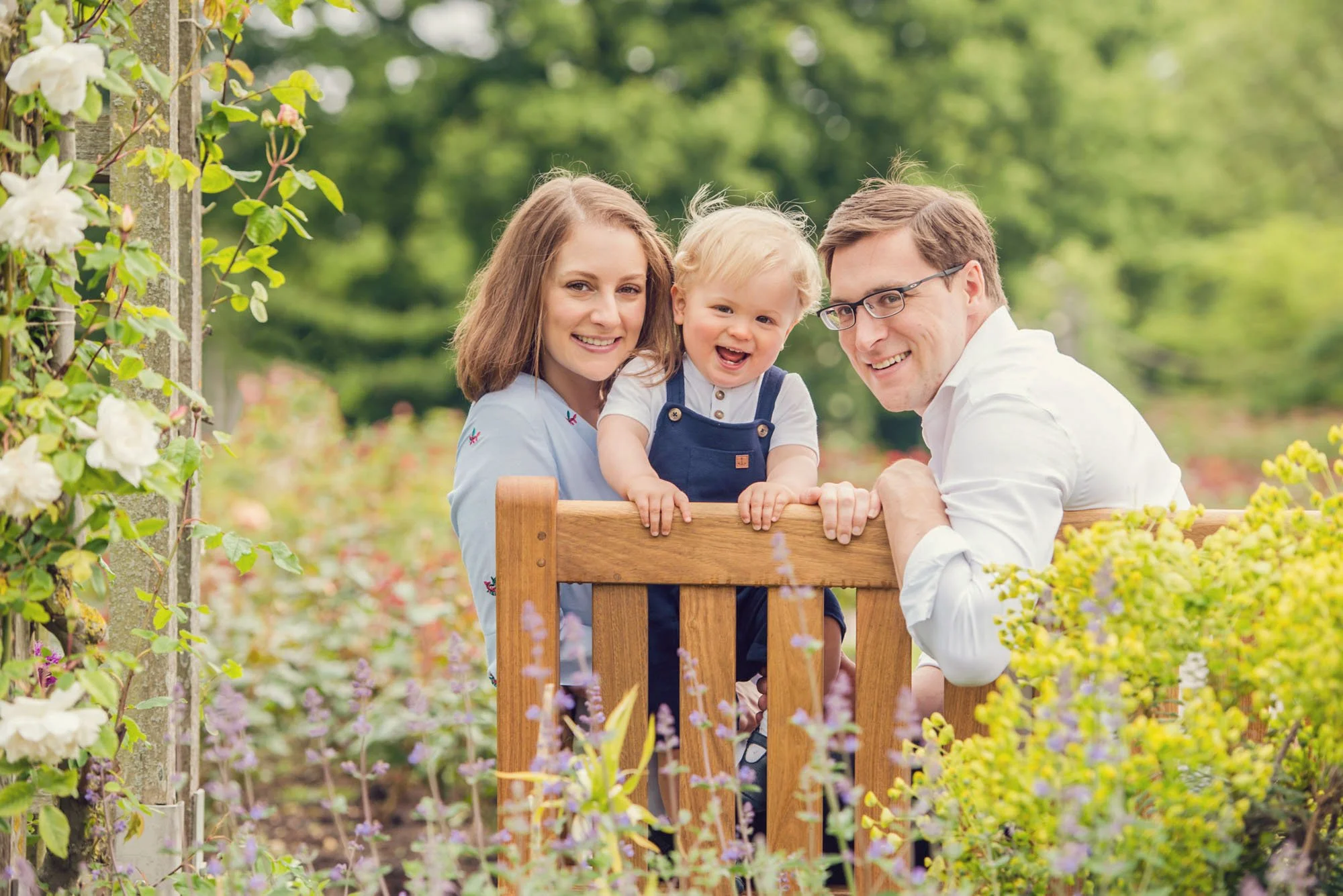 Little boy stands up on a bench between his parents and smiles delightedly at the photographer, surrounded by roses and other flowers in Regent's Park in London