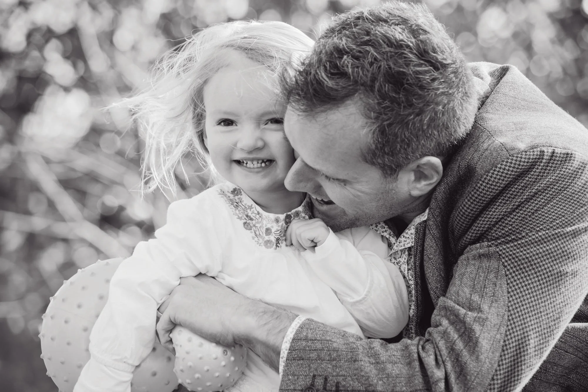 Little girl smiles in delight as he father hugs her tightly during a family photoshoot in Hampstead Heath in north London 