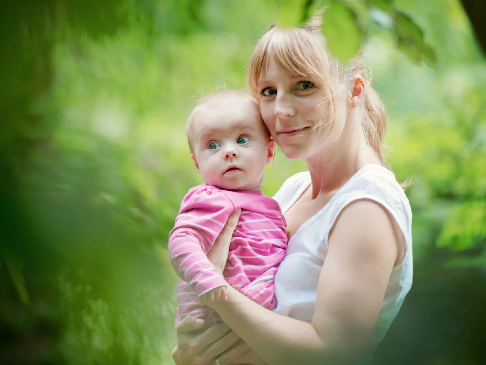 A mother holds her six month old baby in her arms surrounded by green foliage during a summer family photoshoot in Hyde Park in London