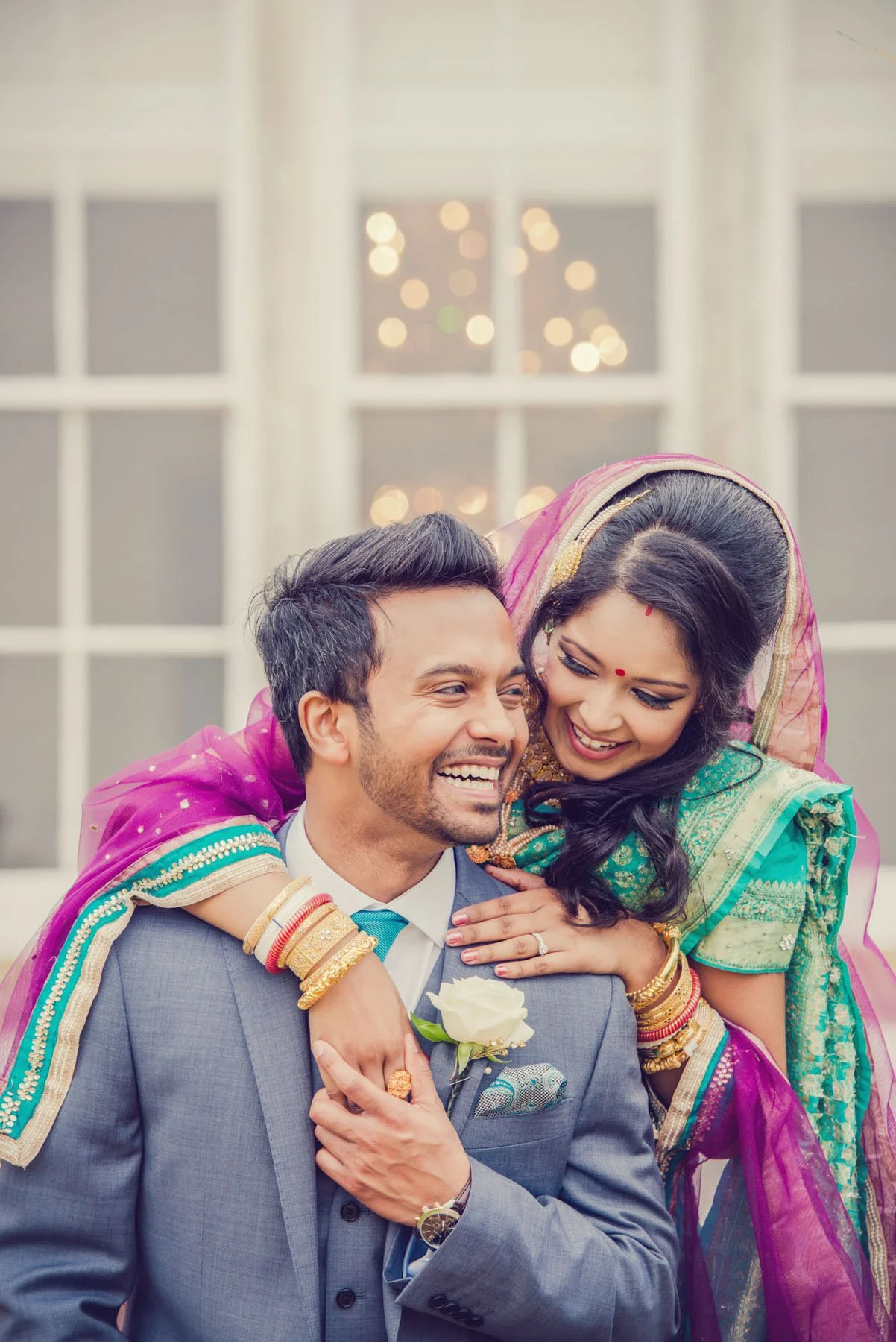 A smiling bride, in a traditional asian wedding outfit, stands behind her laughing groom and hugs him after their weekday wedding at Kenwood House in Hampstead Heath in north London