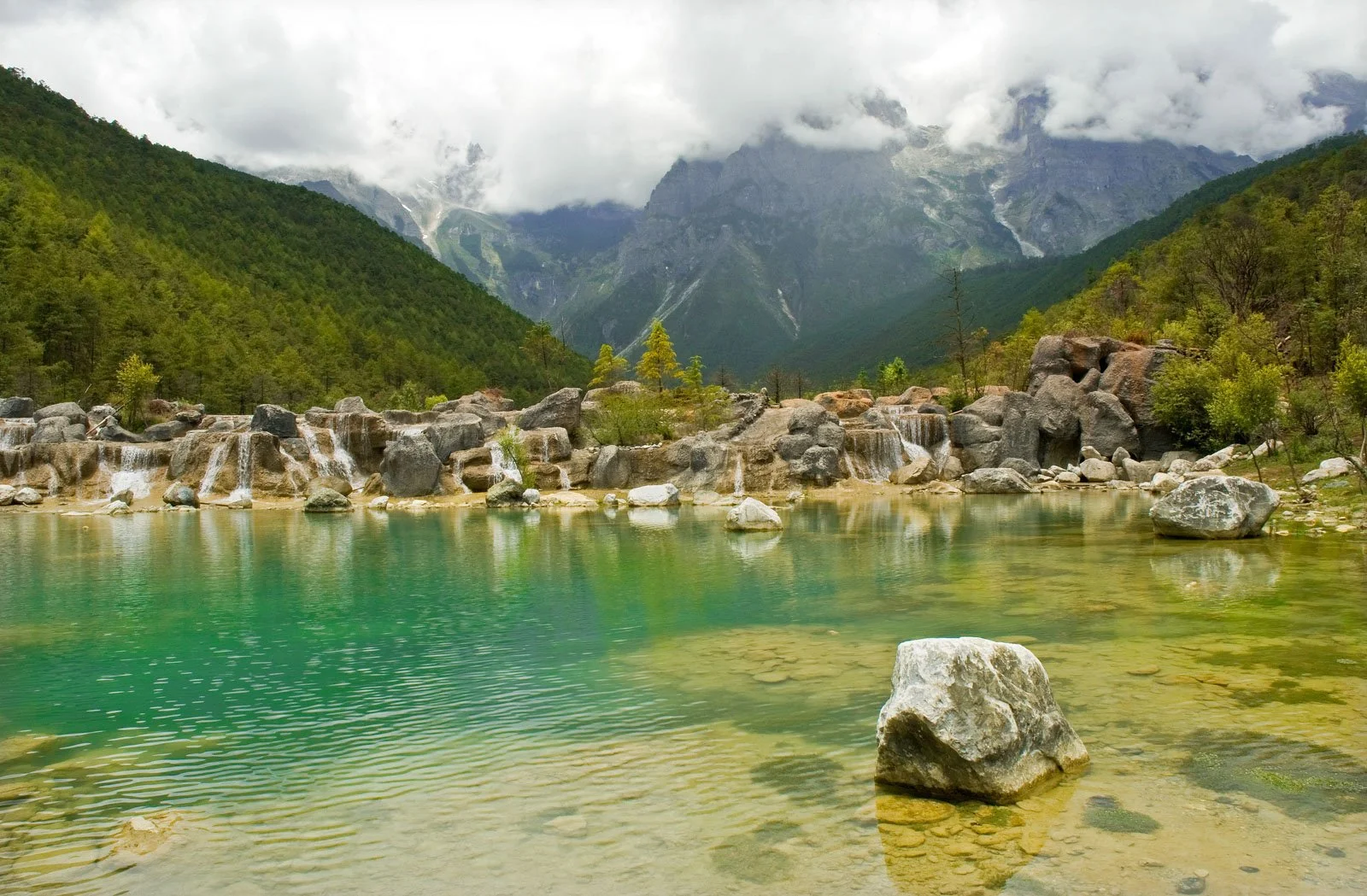 A serene mountain landscape with a clear green lake surrounded by rocks and lush green trees, with misty mountains in the background and cloudy skies.