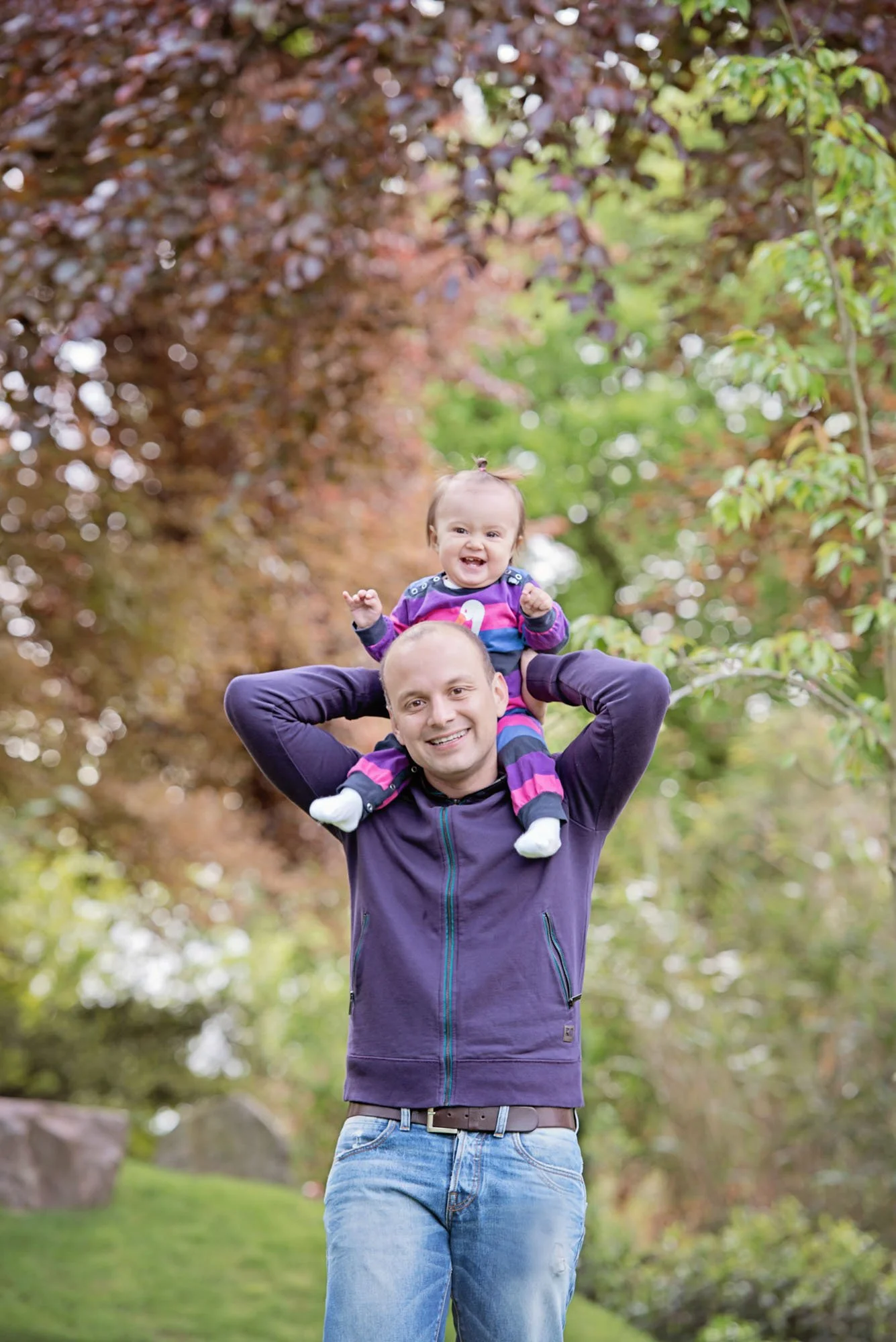 A father holds his laughing one year old baby on his shoulders during a family photoshoot in Holland Park, surrounded by green and red leaves.