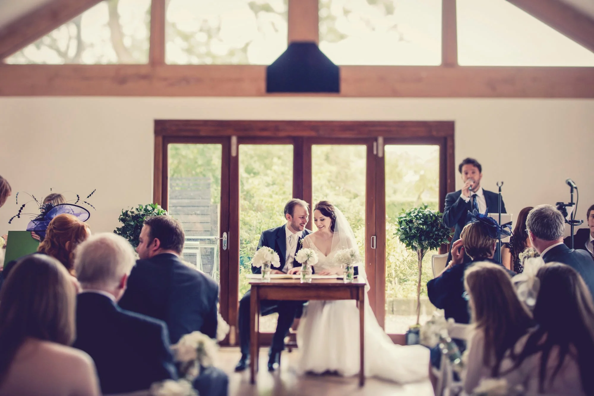 A groom gazes lovingly at his bride as they sign the register during their weekday wedding ceremony at Maidens Barn in Essex