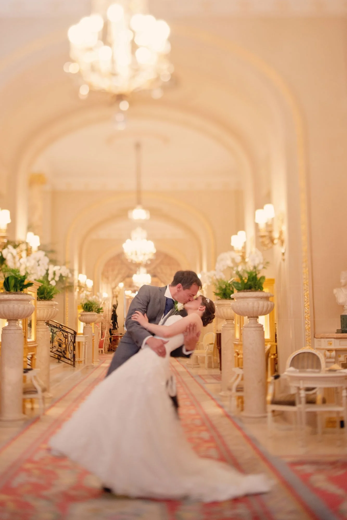 A bride and groom share a passionate kiss under chandeliers in the grand hall of the Ritz Hotel in London