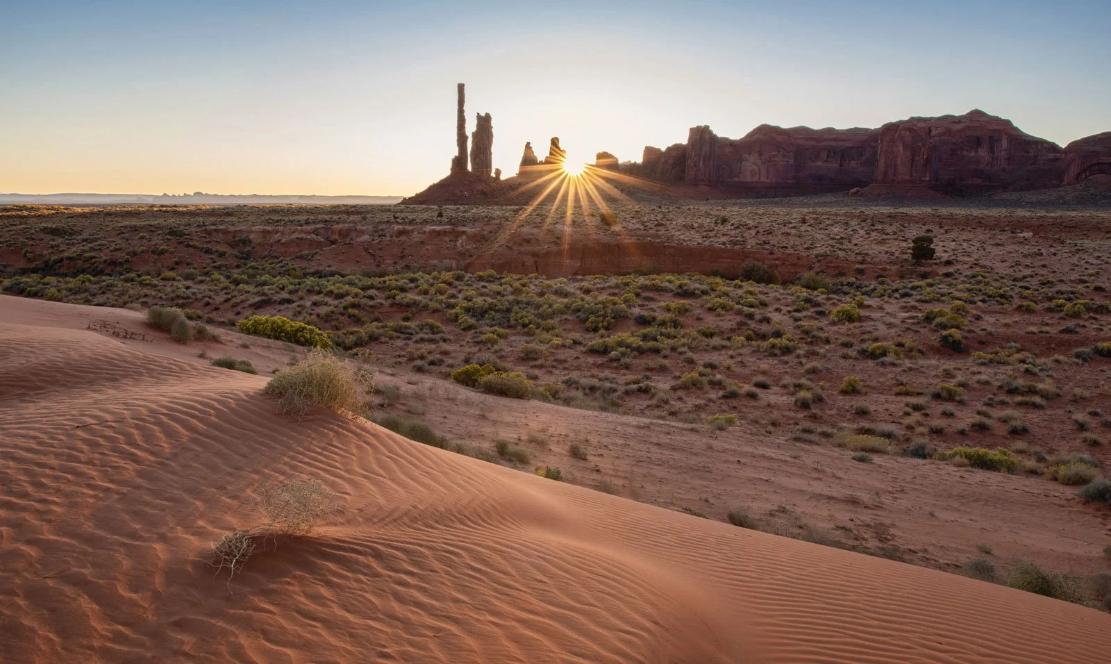 Sandy desert with sparse vegetation in foreground, large rock formations with a sunrise in the background, clear sky.
