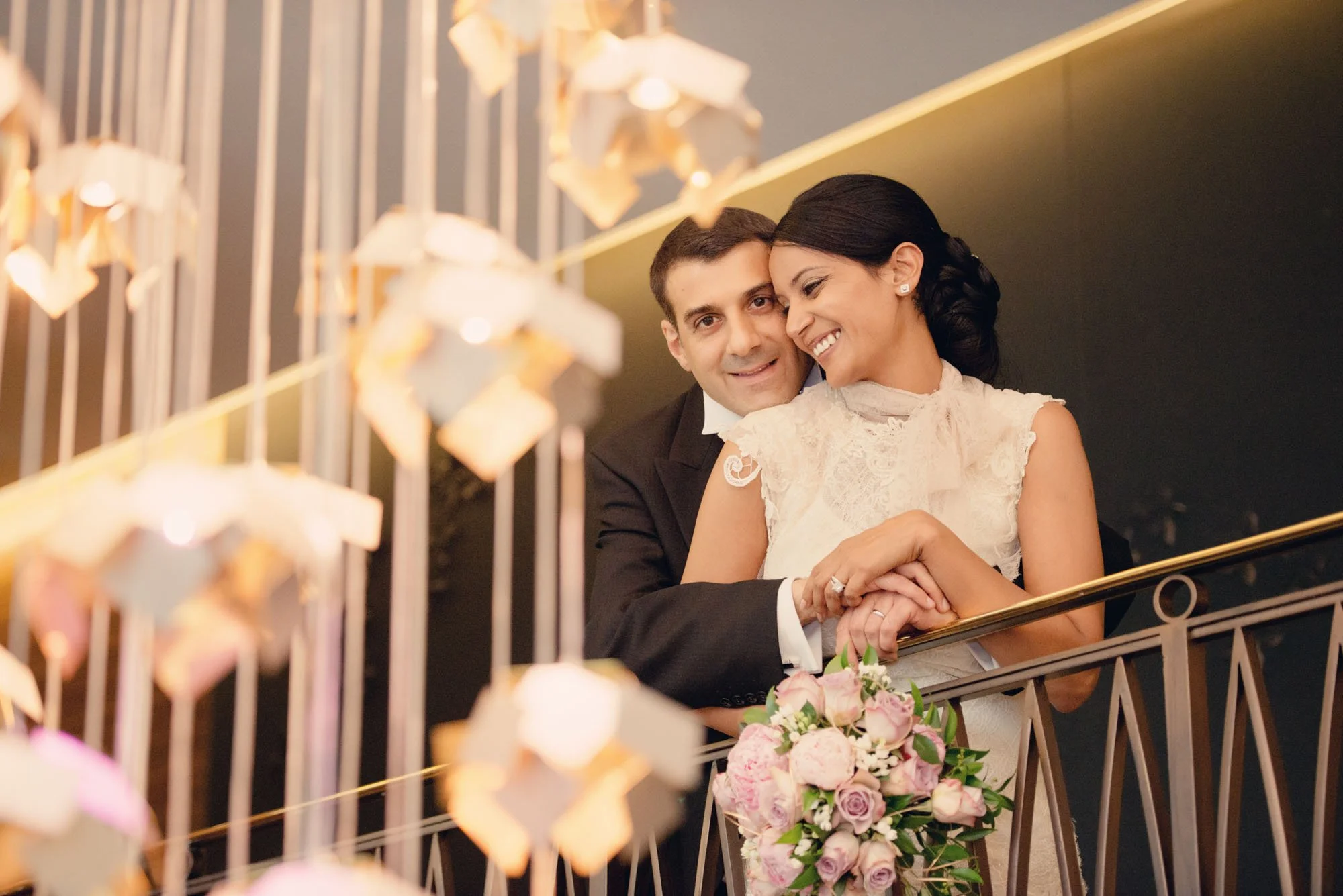 A bride and groom smiling and embracing on a staircase during their wedding at The Langham Hotel in London, with a modern chandelier in front of them.