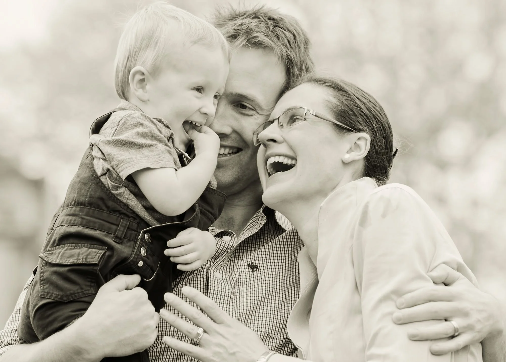 A little boy is hugged by his parents and they all laugh together during a family photoshoot in Hampstead Heath in north London