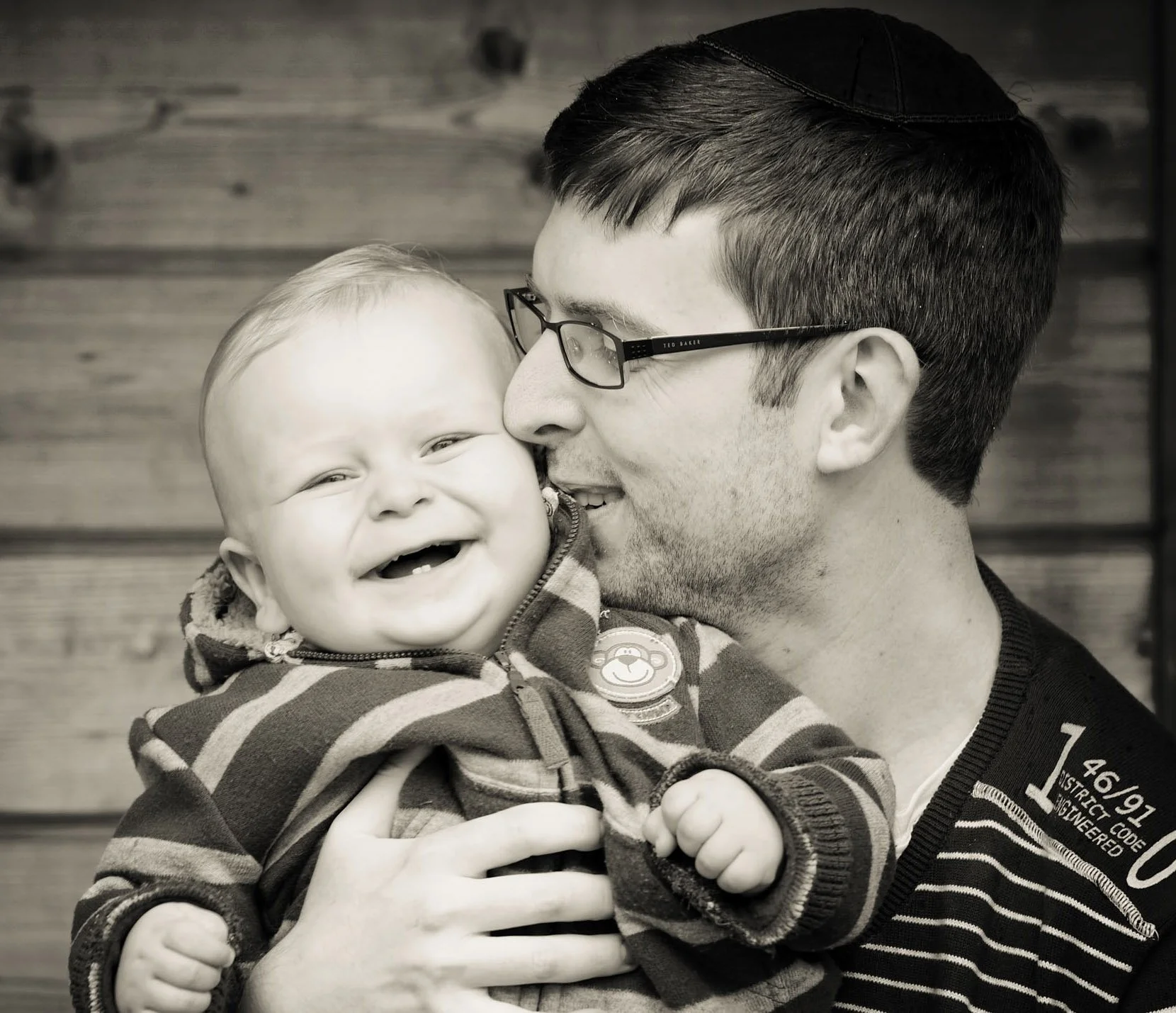 A black and white photo of a father hugging his one year old baby to him as they stand in front of their garden shed at their home in Golders Green in north west London