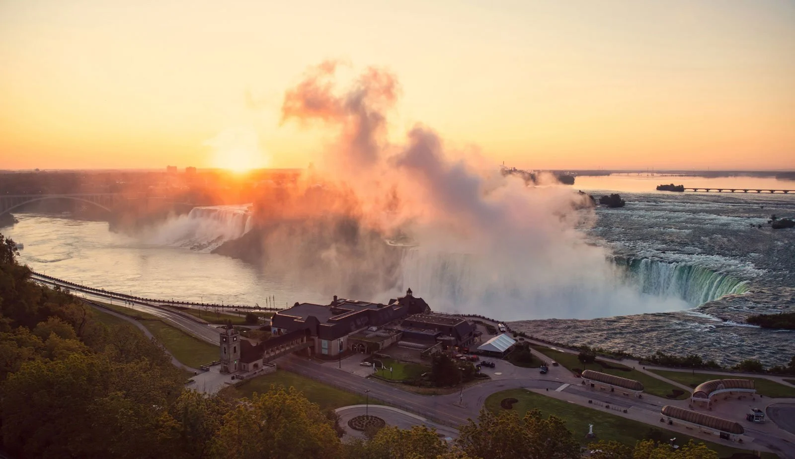 Sunset view of Niagara Falls with mist rising from the waterfalls, a building and trees in the foreground, and a bridge in the background.