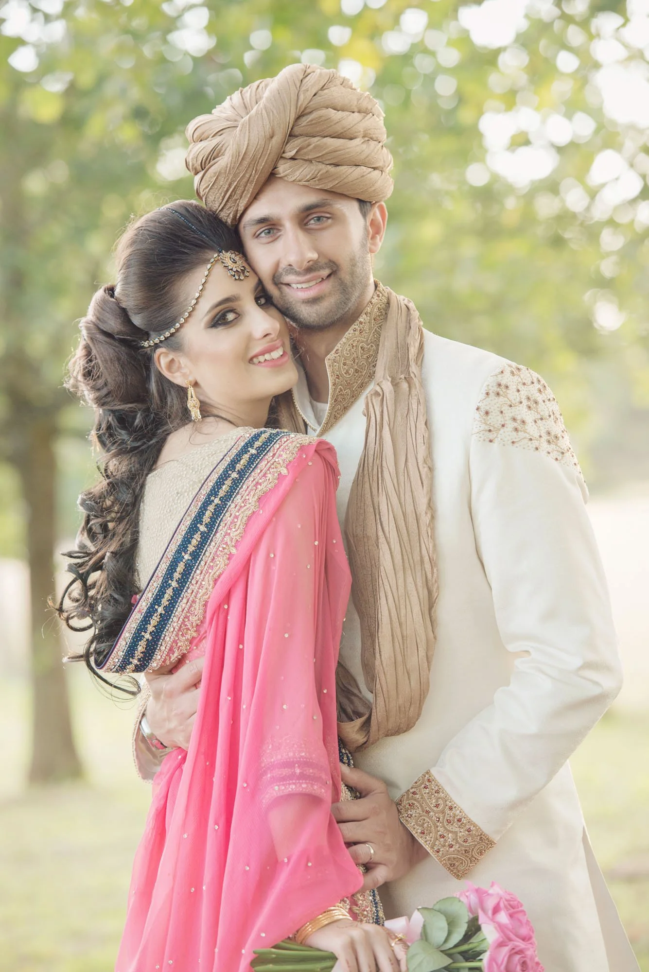 A groom in his asian wedding outfit including turban hugs his asian bride to him as they stand under trees in the grounds of Froyle Park in Hampshire 