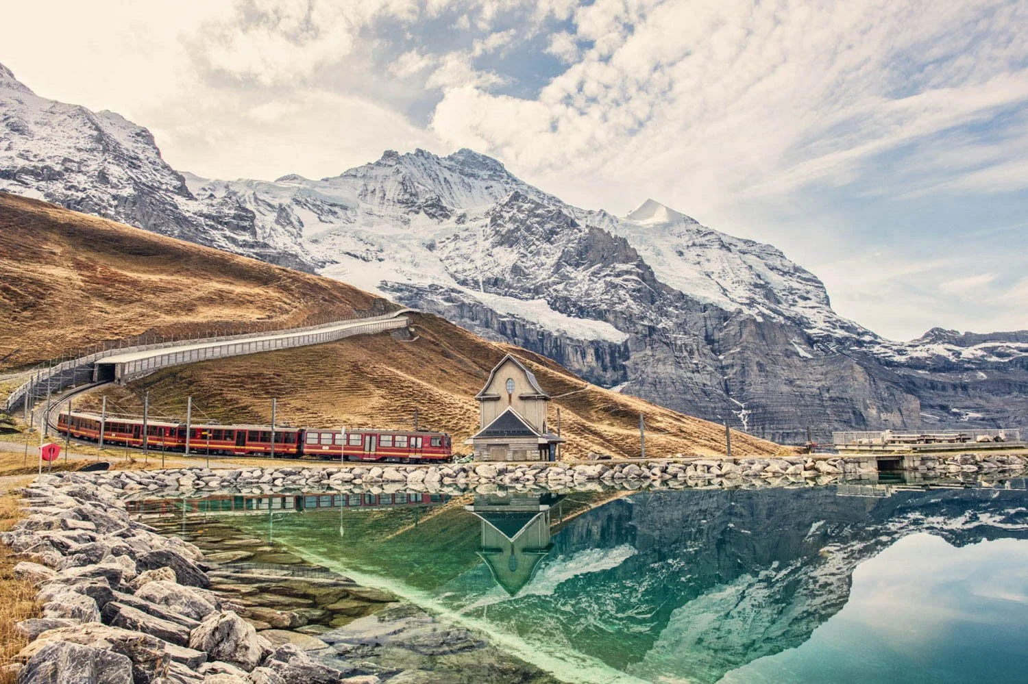 A train is traveling along a curved track beside a lake with mountain backdrop, a small church, rocky shoreline, and snow-capped peaks.