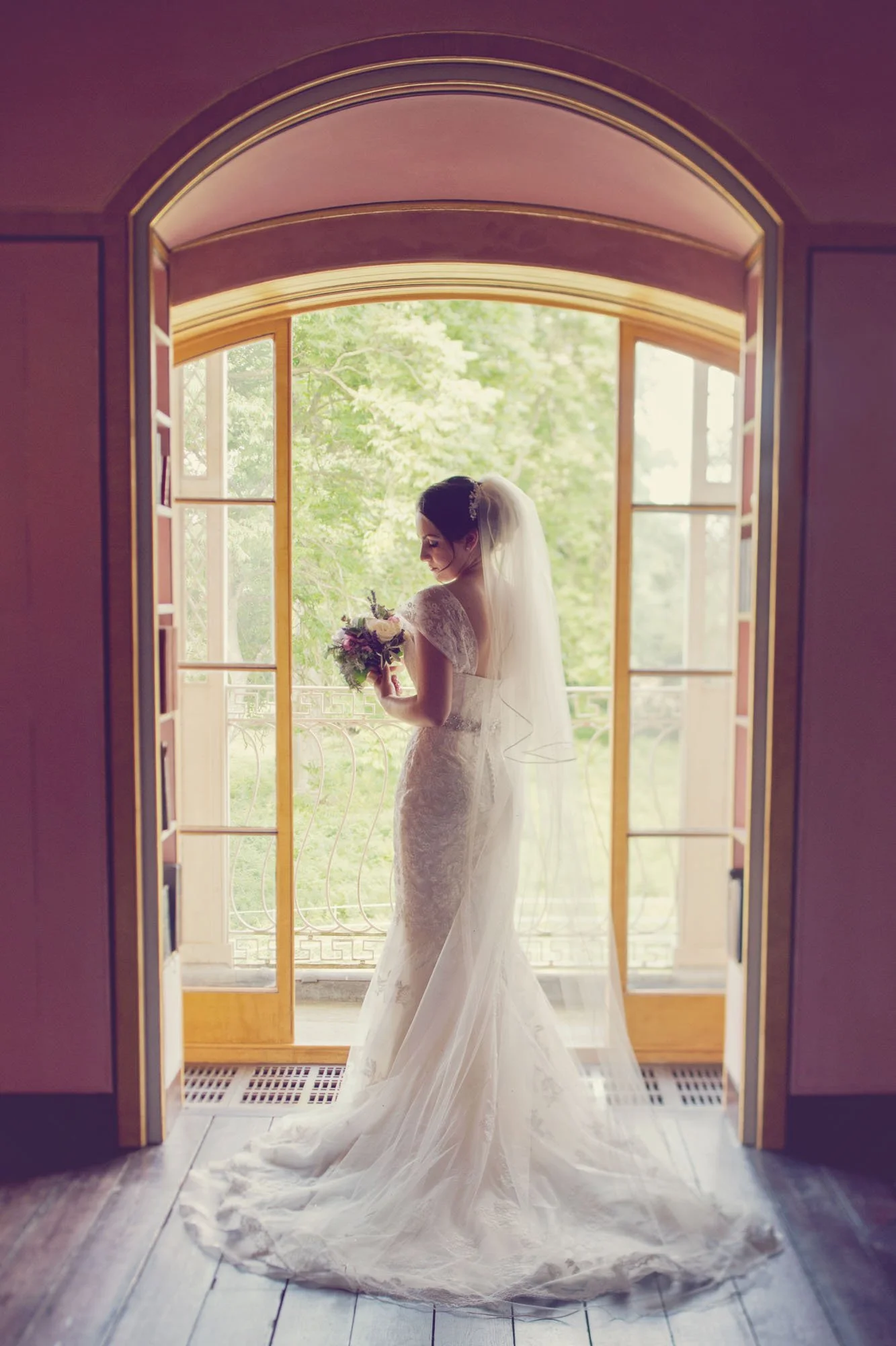 A bride stands in front of a full length window gazing at her bouquet during a micro wedding at Moggerhanger House in Bedfordshire