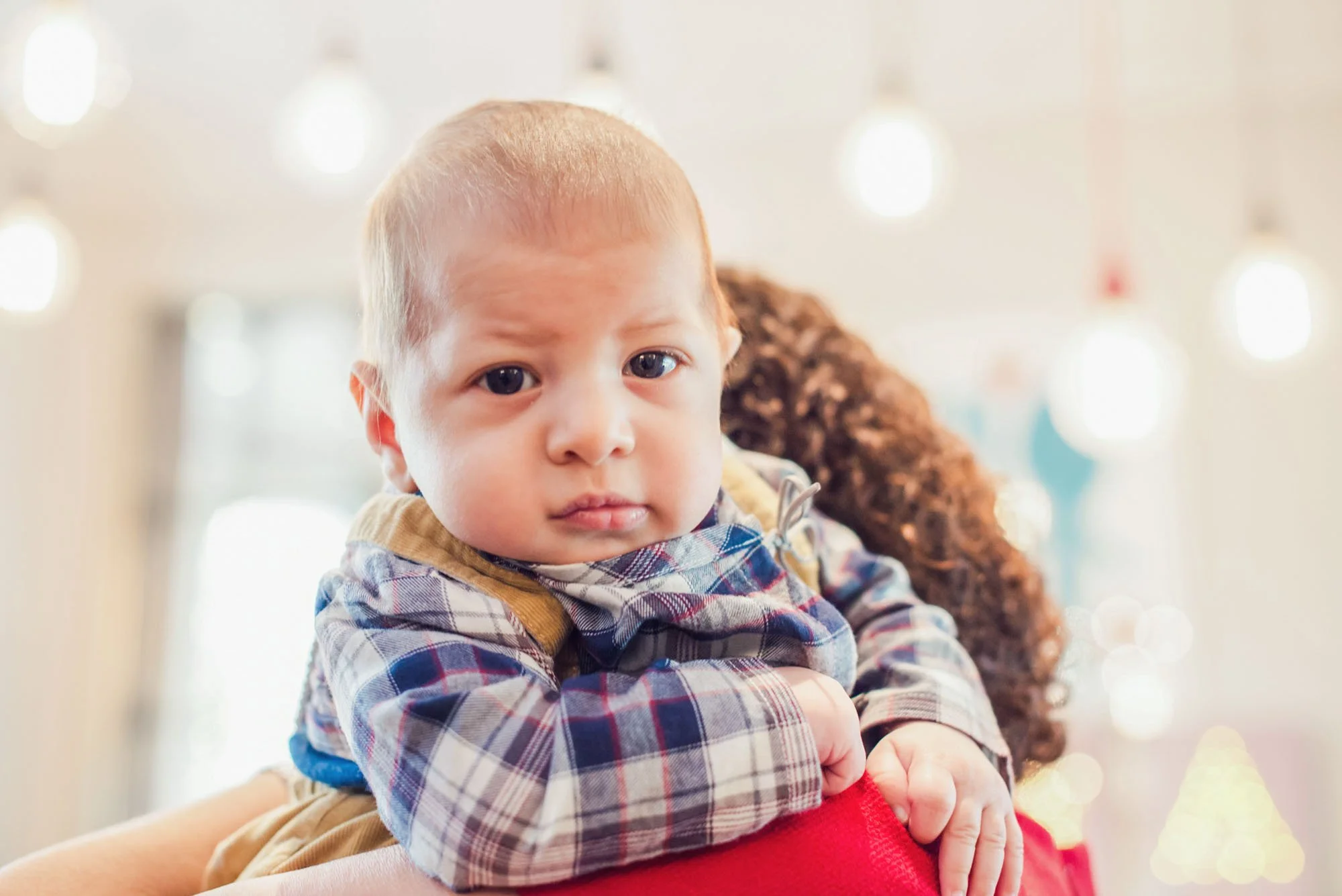 A baby boy props himself up on his elbows as he's held on his mother's shoulder during a family photoshoot in Kentish Town in north London.  He is wearing a checked shirt and looks directly into the camera.