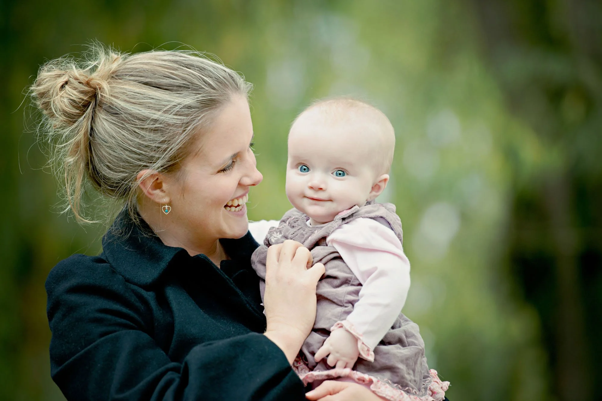A mother smiles at her six month old baby as she holds the baby in her arms in front of green leaves in Hyde Park in London
