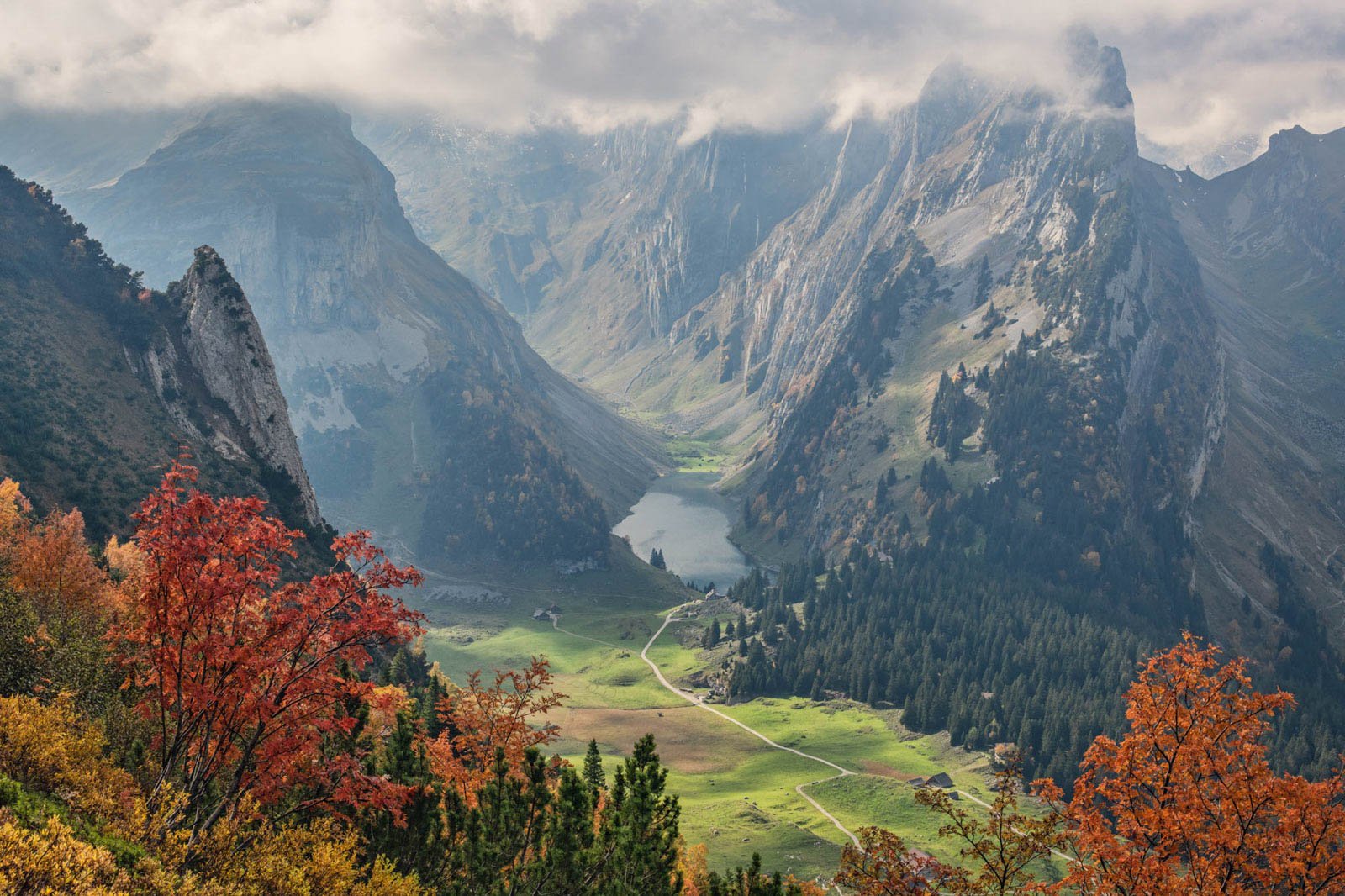 A scenic mountain landscape with tall, rugged peaks in the background, a valley with a winding path, a small lake, and colorful autumn trees in the foreground.