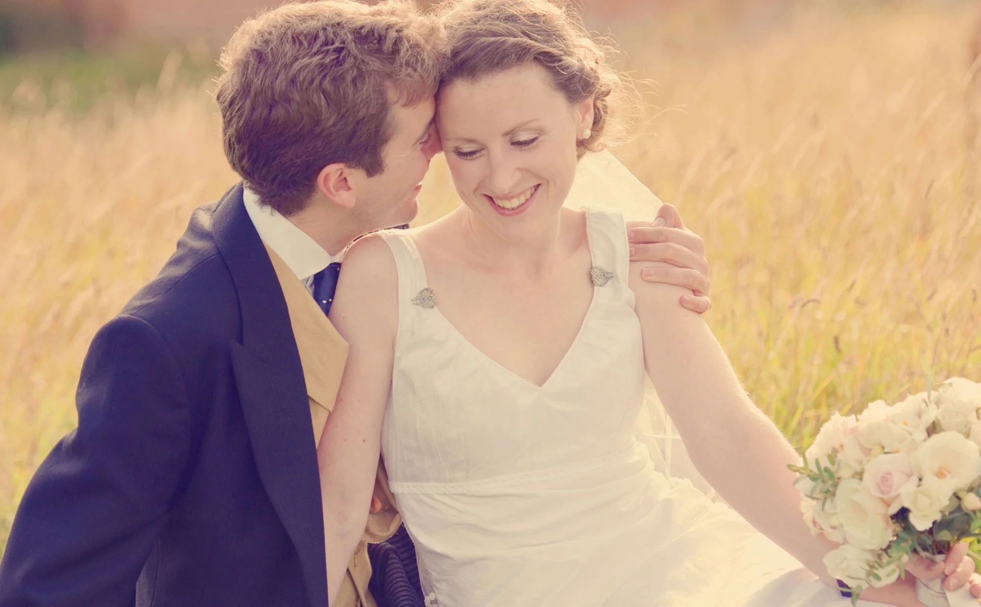 A groom holds his bride as they sit in golden grass in Hampstead Heath after their wedding at Burgh House in Hampstead
