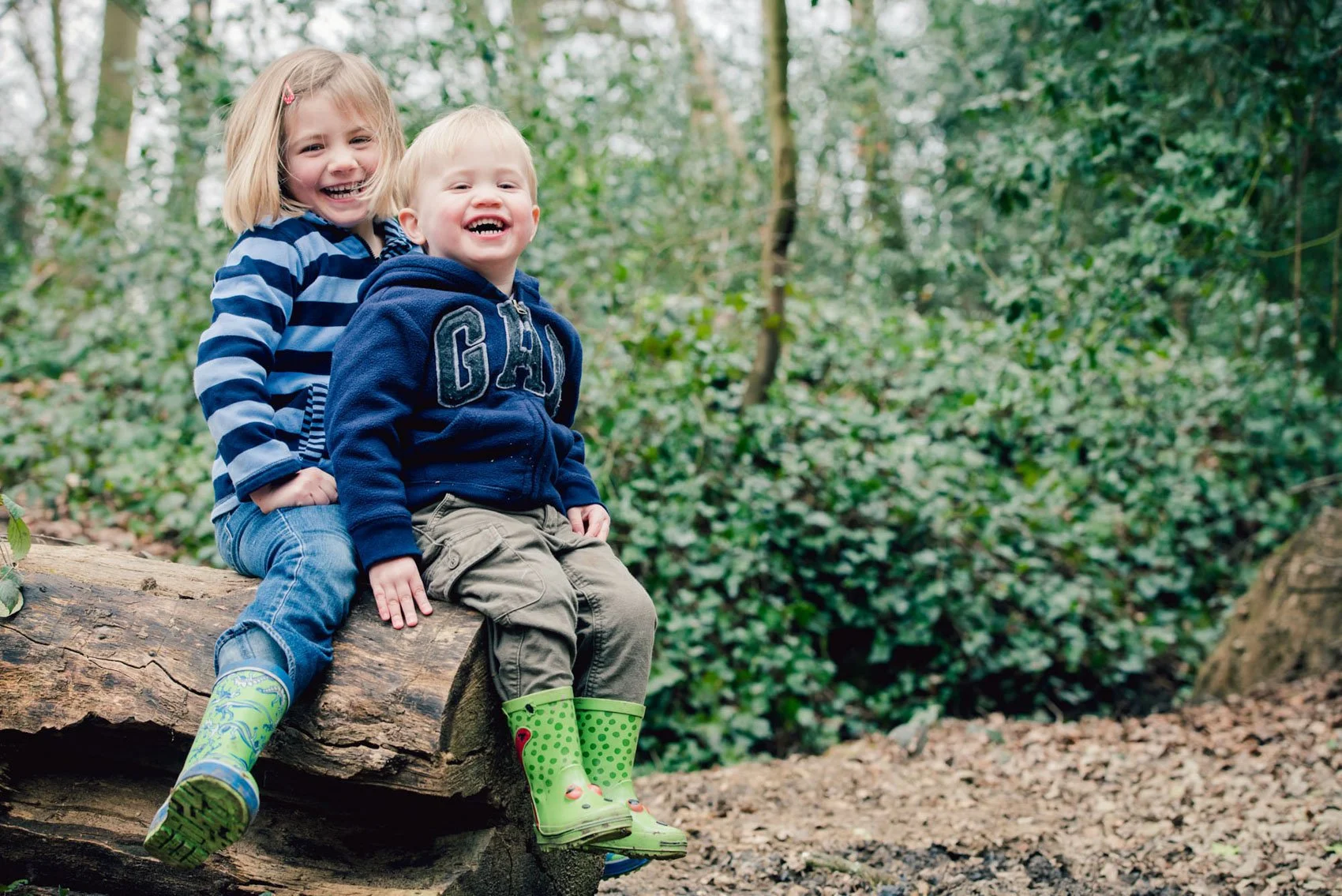 A young brother and sister wearing wellies sit on a log during a family photoshoot in Cannizaro Park in Wimbledon in south west London 