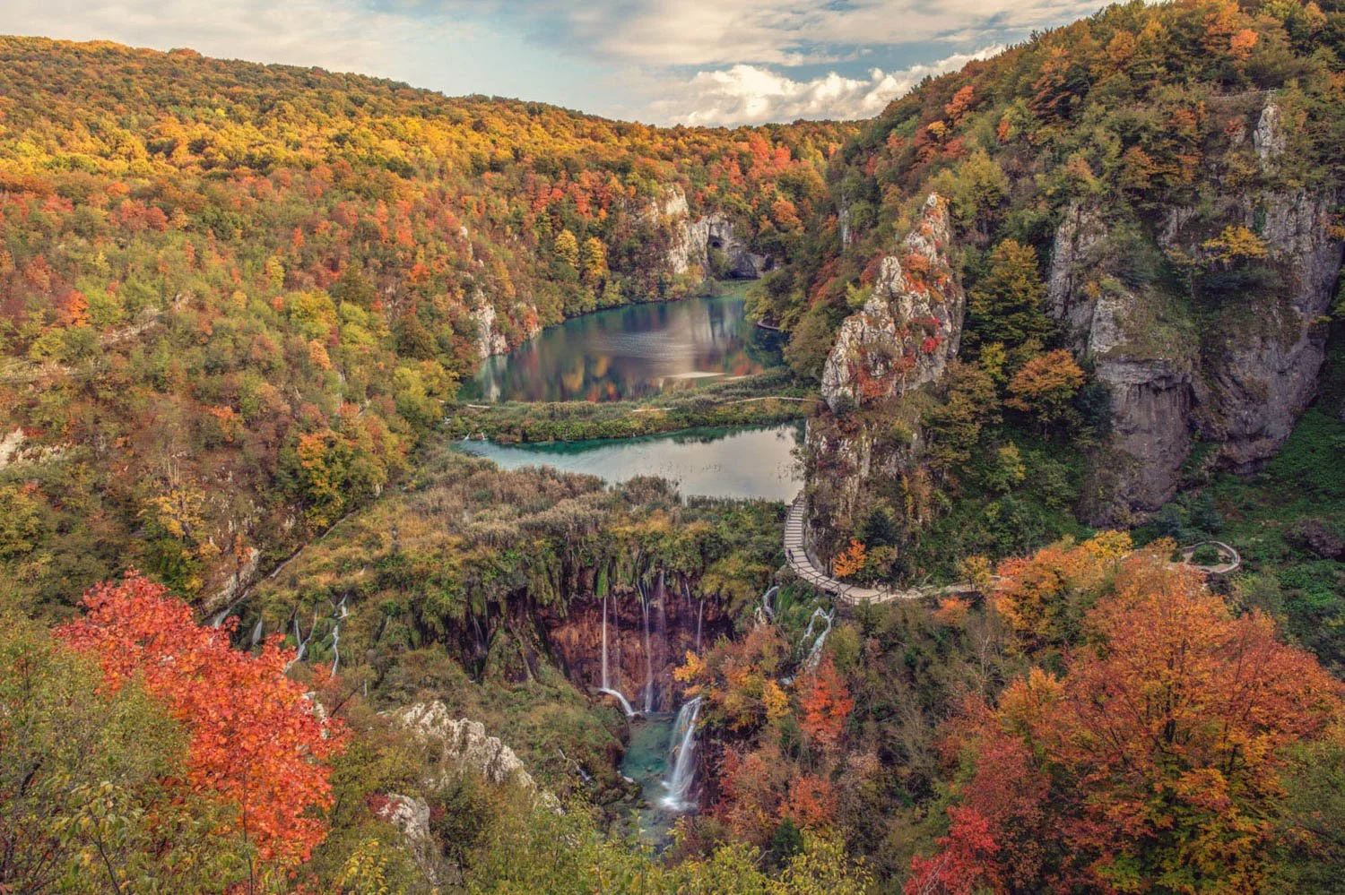 A scenic landscape of a canyon with colorful autumn foliage, two lakes, waterfalls, and a winding pathway along the cliffs.