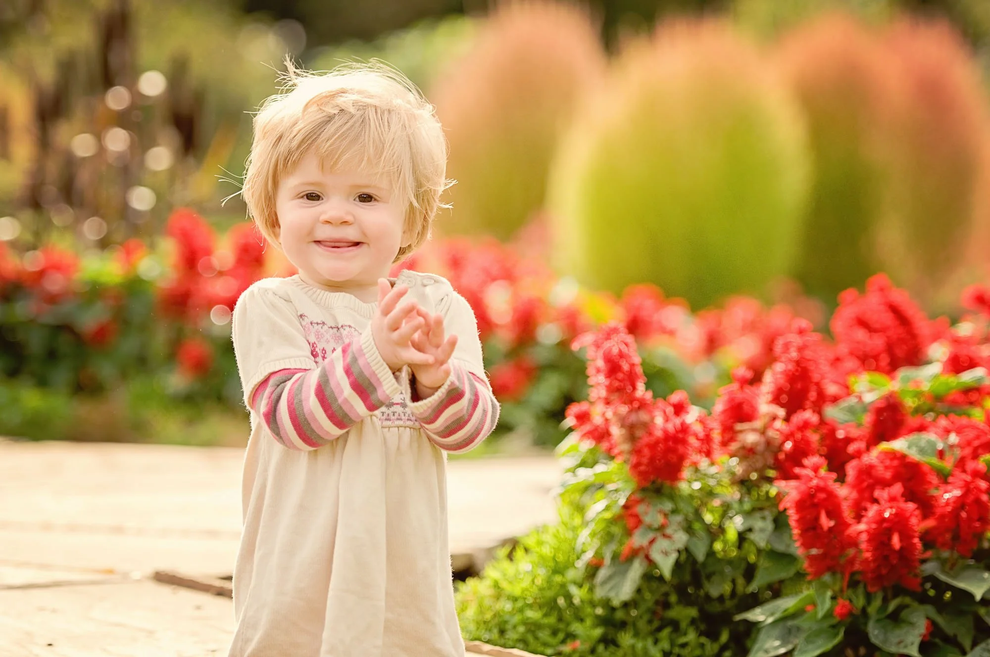 A little girl claps her hands together and smiles as she stands next to red flowers in Cannizaro Park in Wimbledon in south west London
