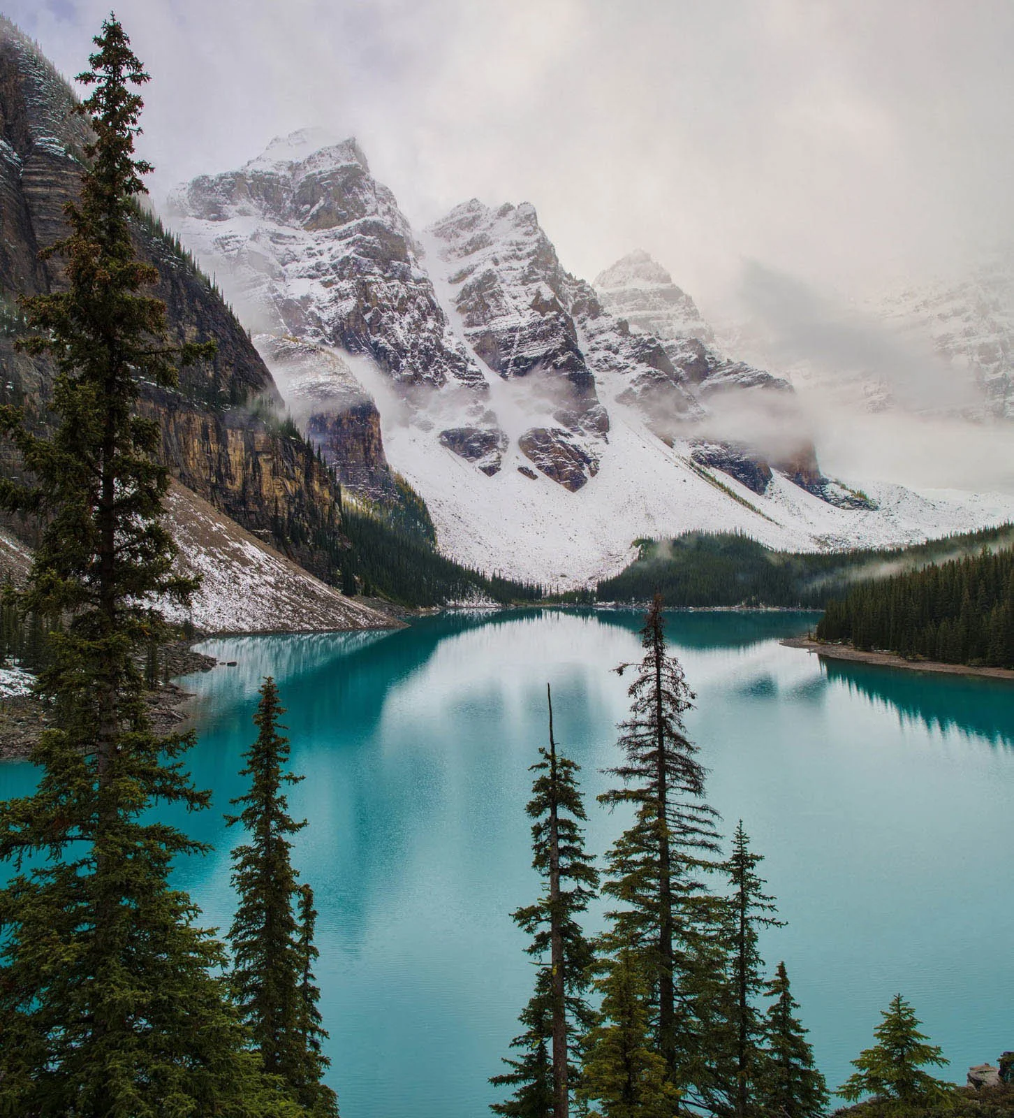 Snow-capped mountains behind a turquoise lake, with pine trees in the foreground and mist around the peaks.