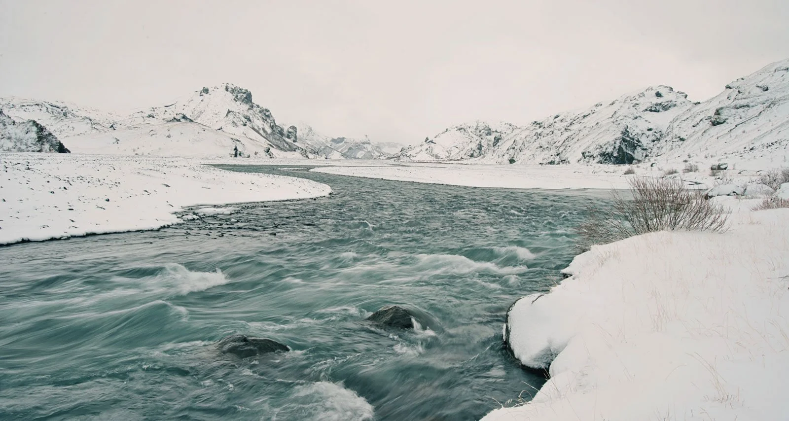 A river flowing through a snowy landscape with snow-covered mountains in the background.