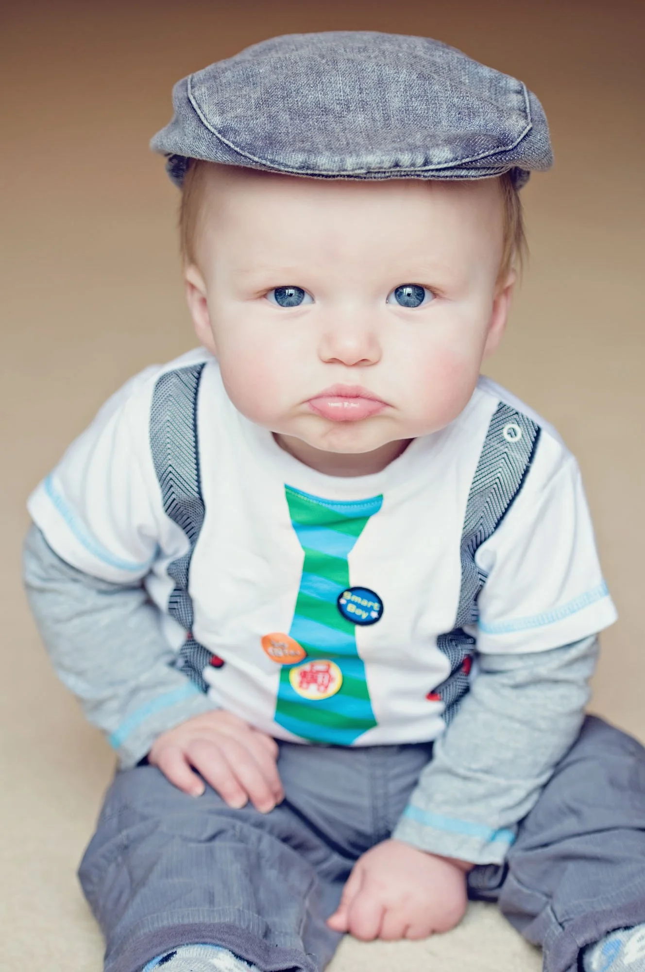 An eight month old baby boy wears a flat cap and pouts up at the camera with a hand on his hip, during a family photoshoot in Ealing in west London