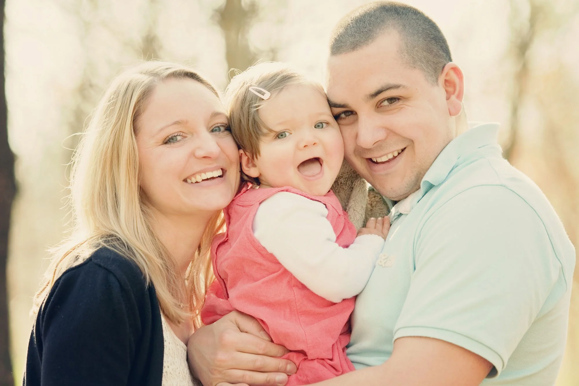 Little girl laughs as she is cuddled by her parents during a family photoshoot in Hampstead Heath in London, with golden light shining through the trees behind them