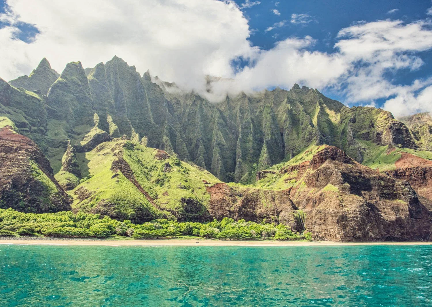 Tropical landscape with turquoise water, green foliage, and tall green mountains with clouds and blue sky.