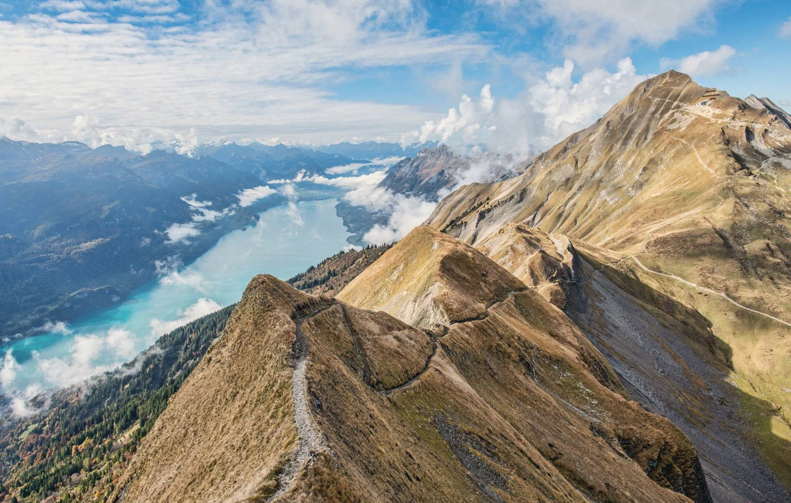 Mountain ridge with a winding trail, overlooking a deep lake with surrounding mountains and a partly cloudy sky.